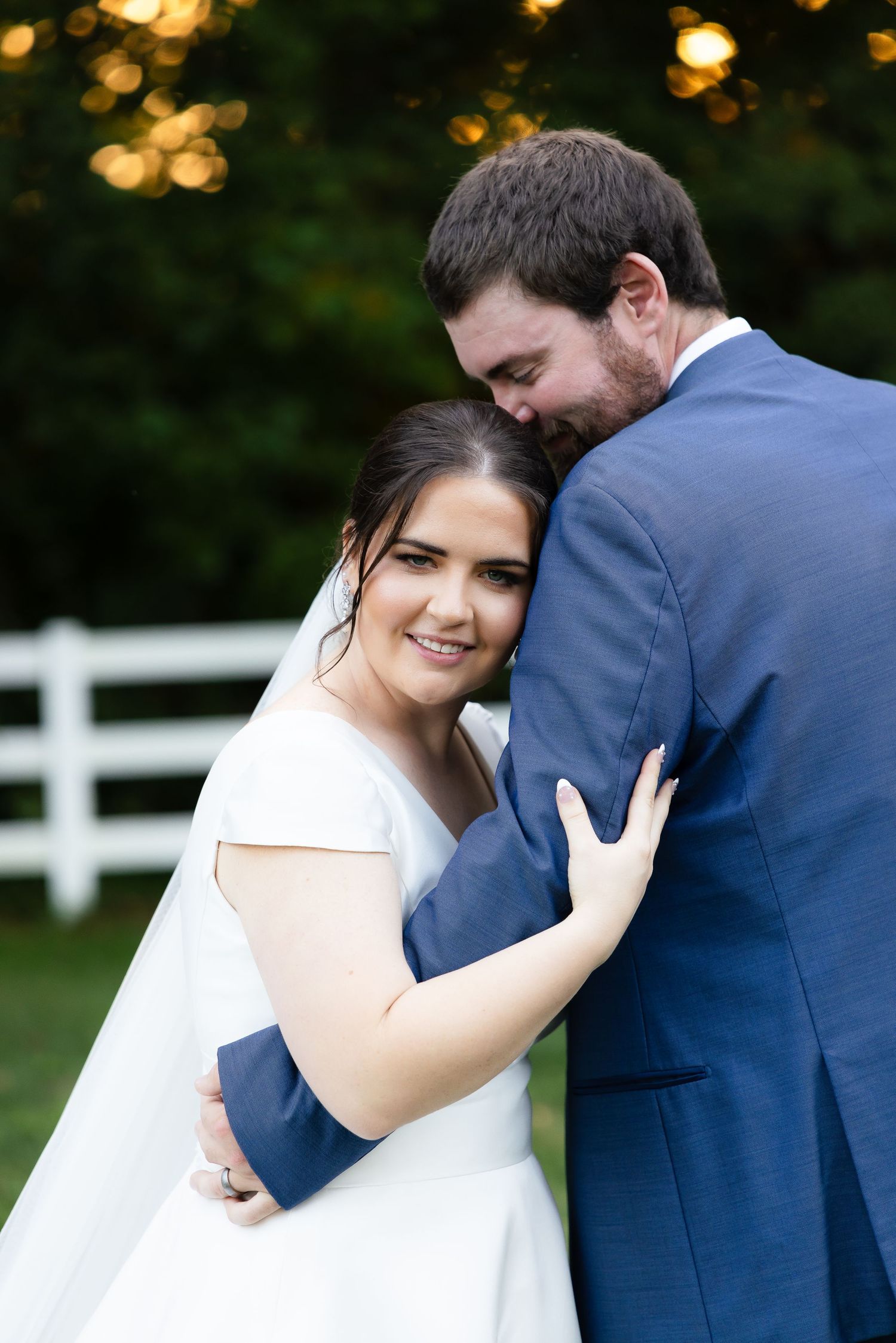 A romantic wedding portrait of a couple embracing outdoors at sunset with a white fence in the background.
