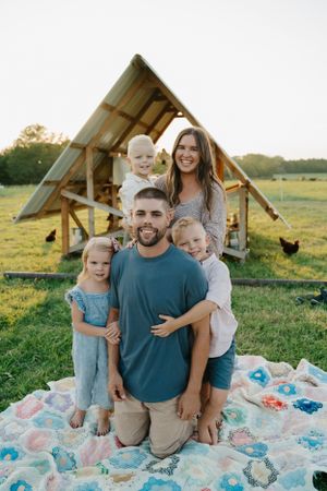 Happy family sitting together on a colorful blanket during golden hour with a wooden shelter behind them.