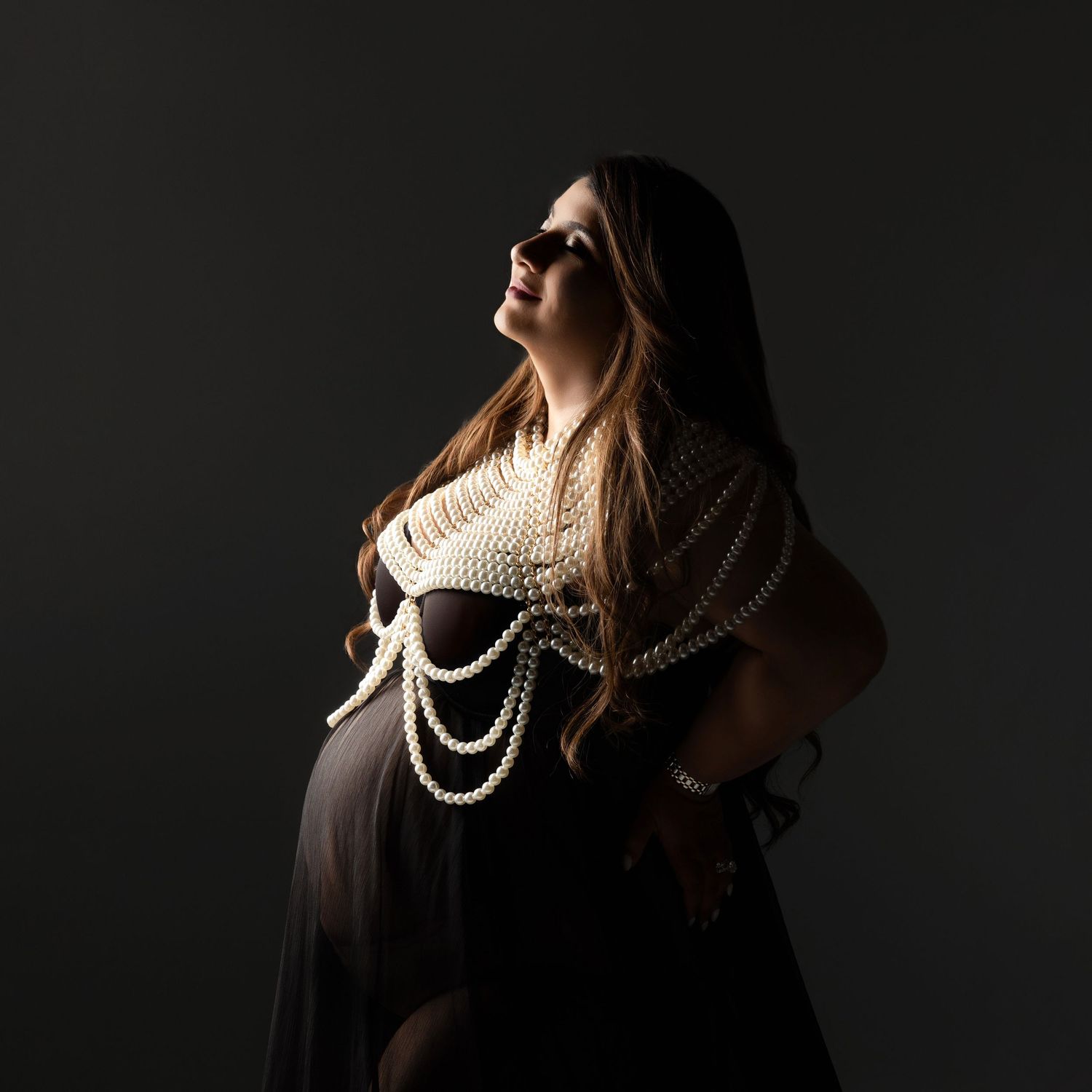 Dramatic portrait in low light with draped pearl necklaces against dark background creating moody artistic effect.