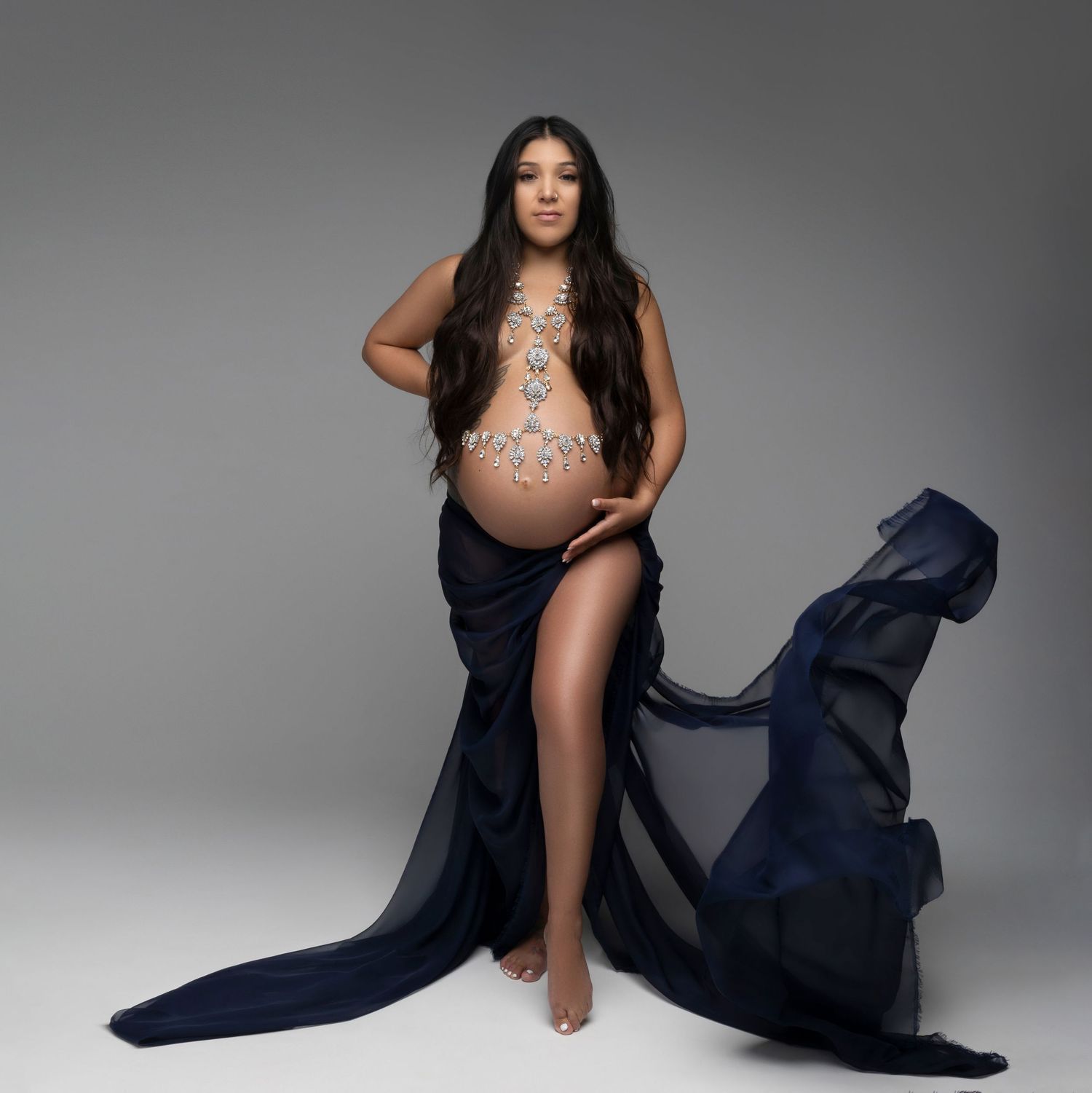 Elegant maternity photo of a woman in a flowing navy dress with a lace bodysuit against a gray studio backdrop.