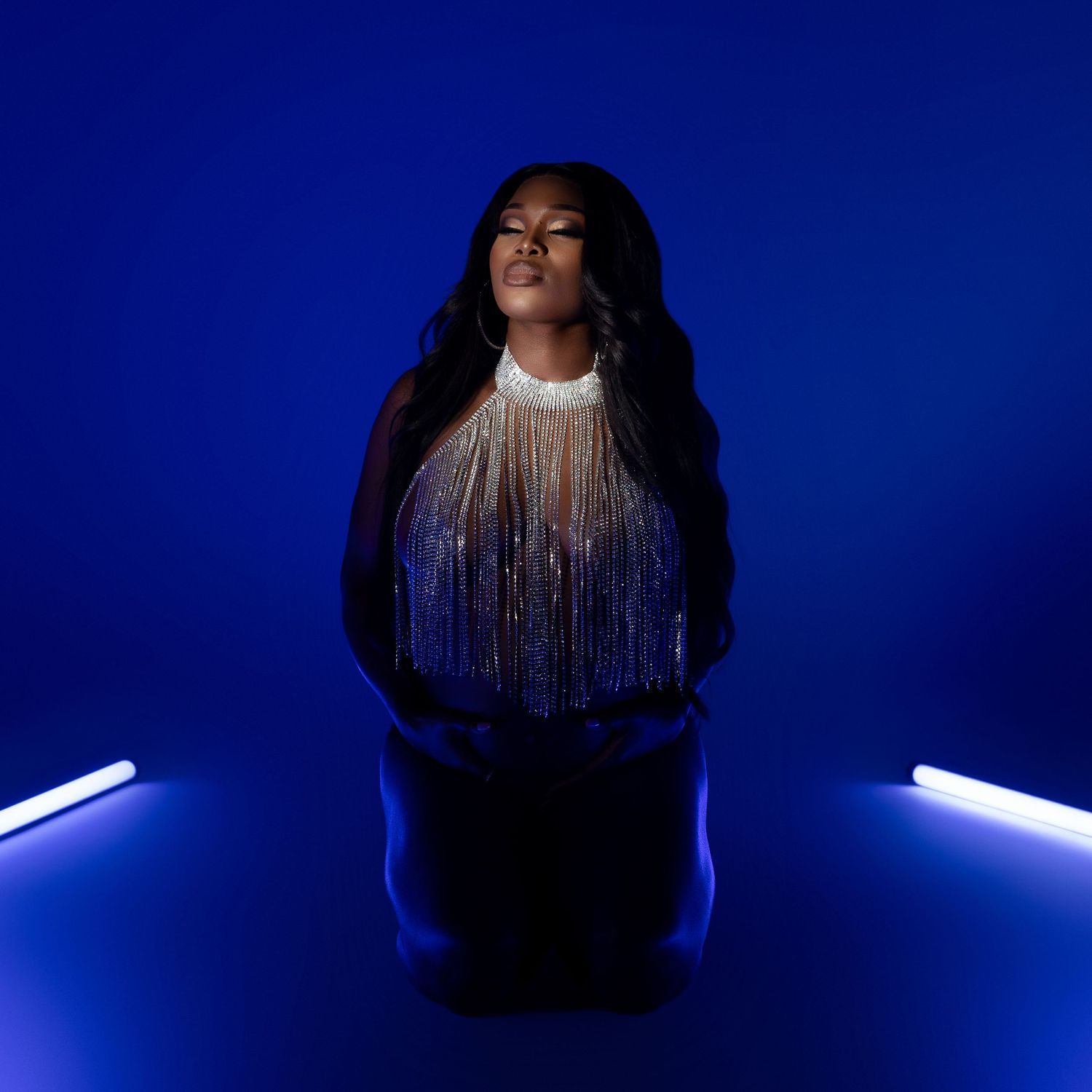 Studio portrait on deep blue backdrop with silver fringe necklace and black top illuminated by neon lights at sides.