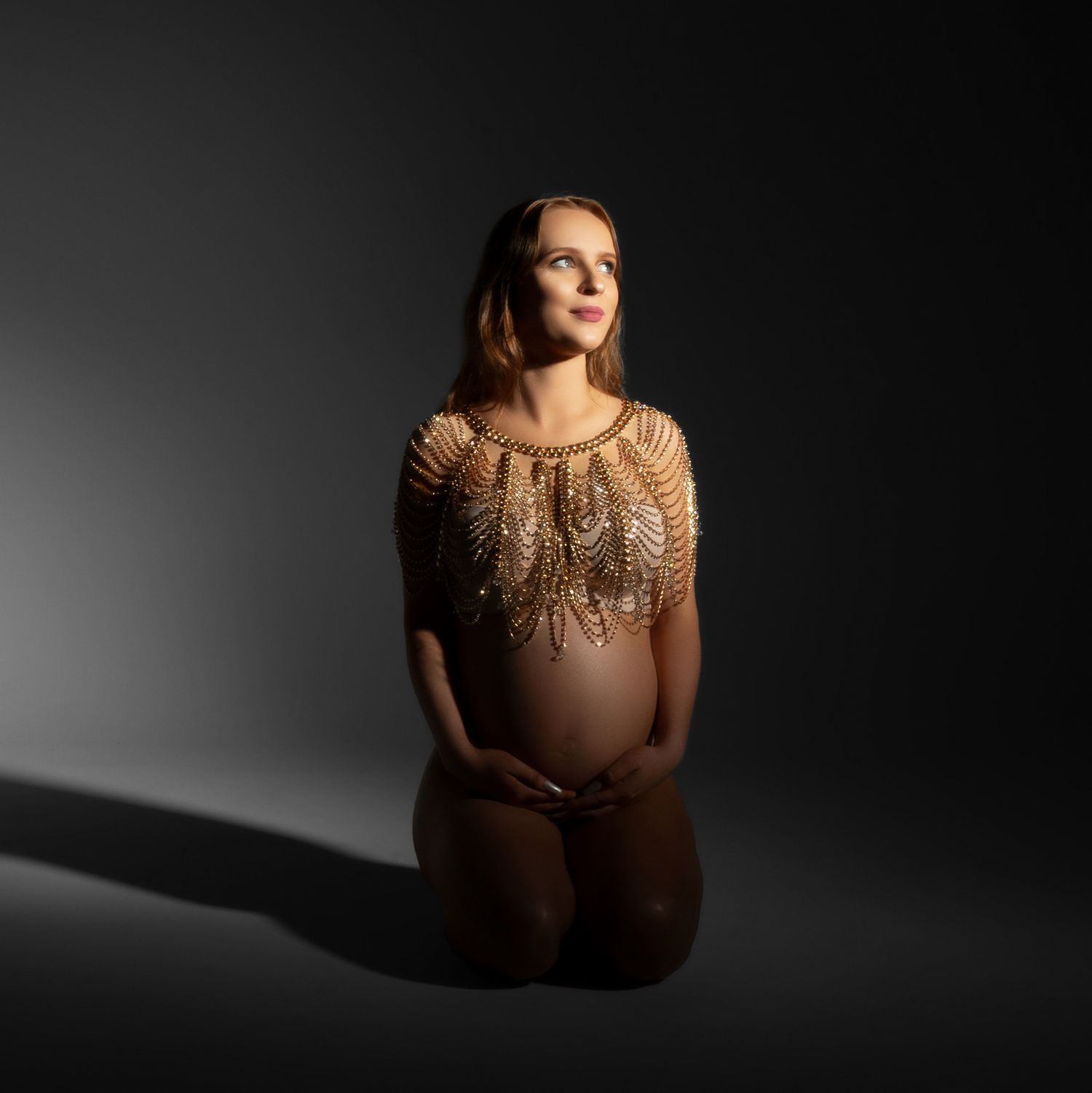 Maternity portrait in dramatic lighting shows expectant mother wearing shimmering gold evening gown against dark background.
