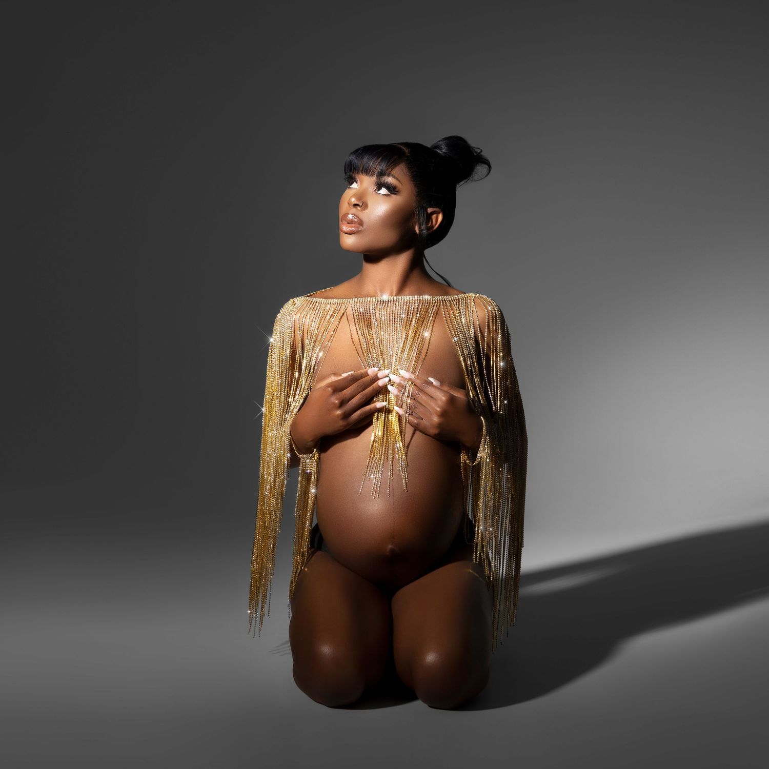 Artistic maternity photo of expectant mother in gold fringe top and black bottoms against gray background.