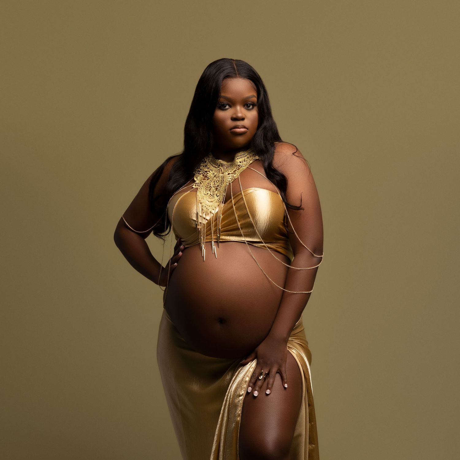 A maternity photo featuring someone wearing a glamorous gold ensemble against a brown backdrop in studio lighting.