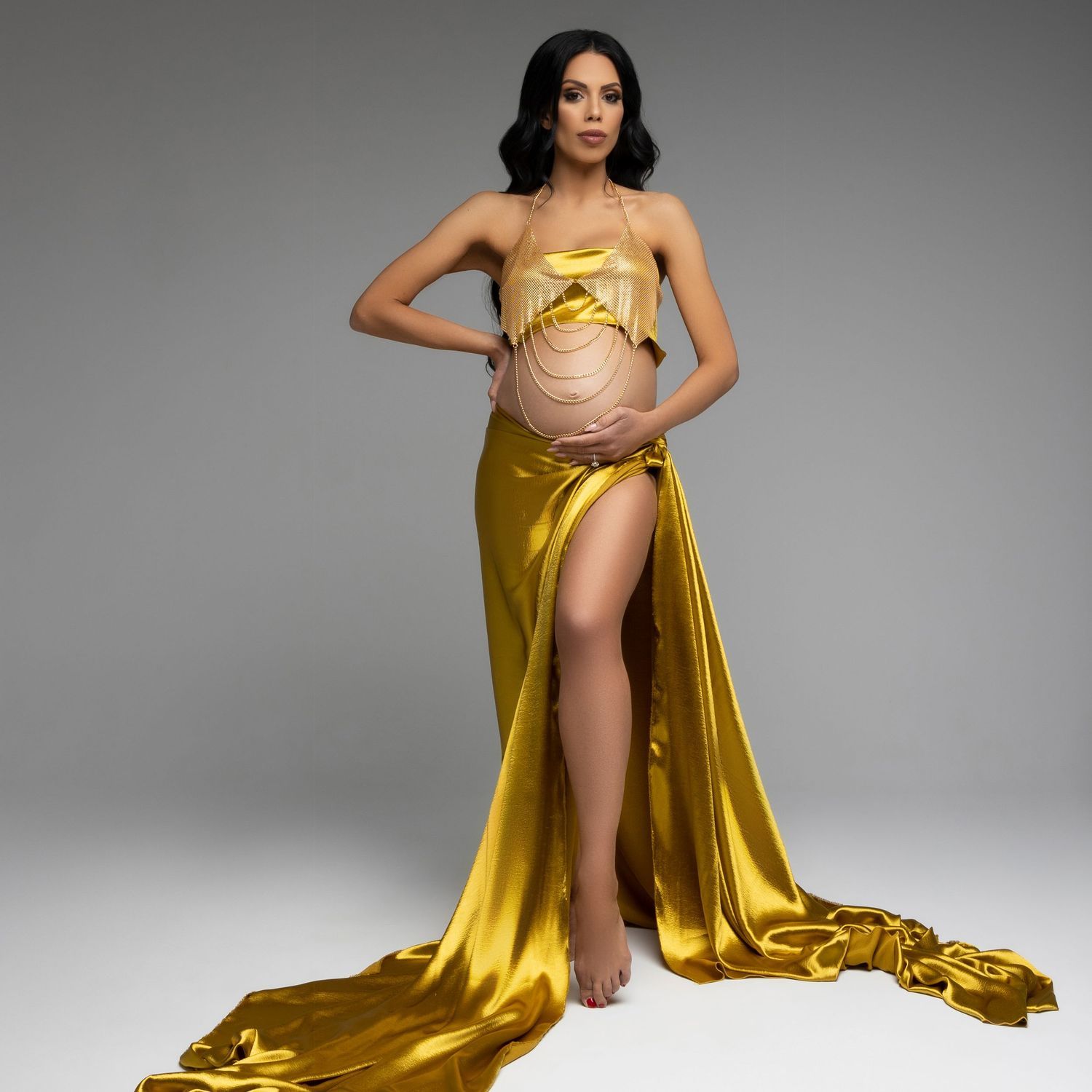 Pregnant woman in gold satin gown with thigh slit poses elegantly against gray backdrop for maternity photoshoot.