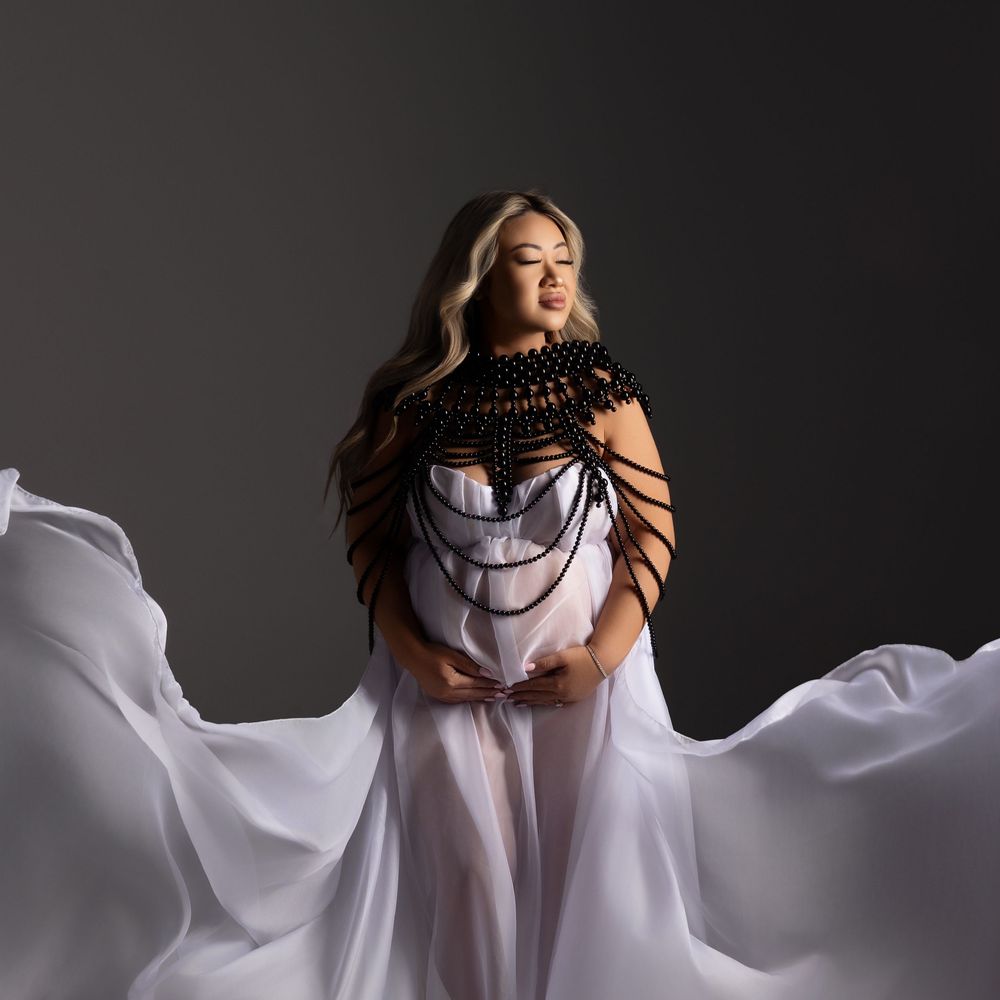A flowing white sheer gown with black lace neckline detail billows dramatically against a dark backdrop in a fashion photo.