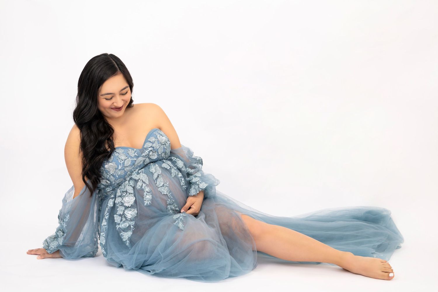 A maternity photo shoot featuring an ethereal light blue tulle gown against a white backdrop.