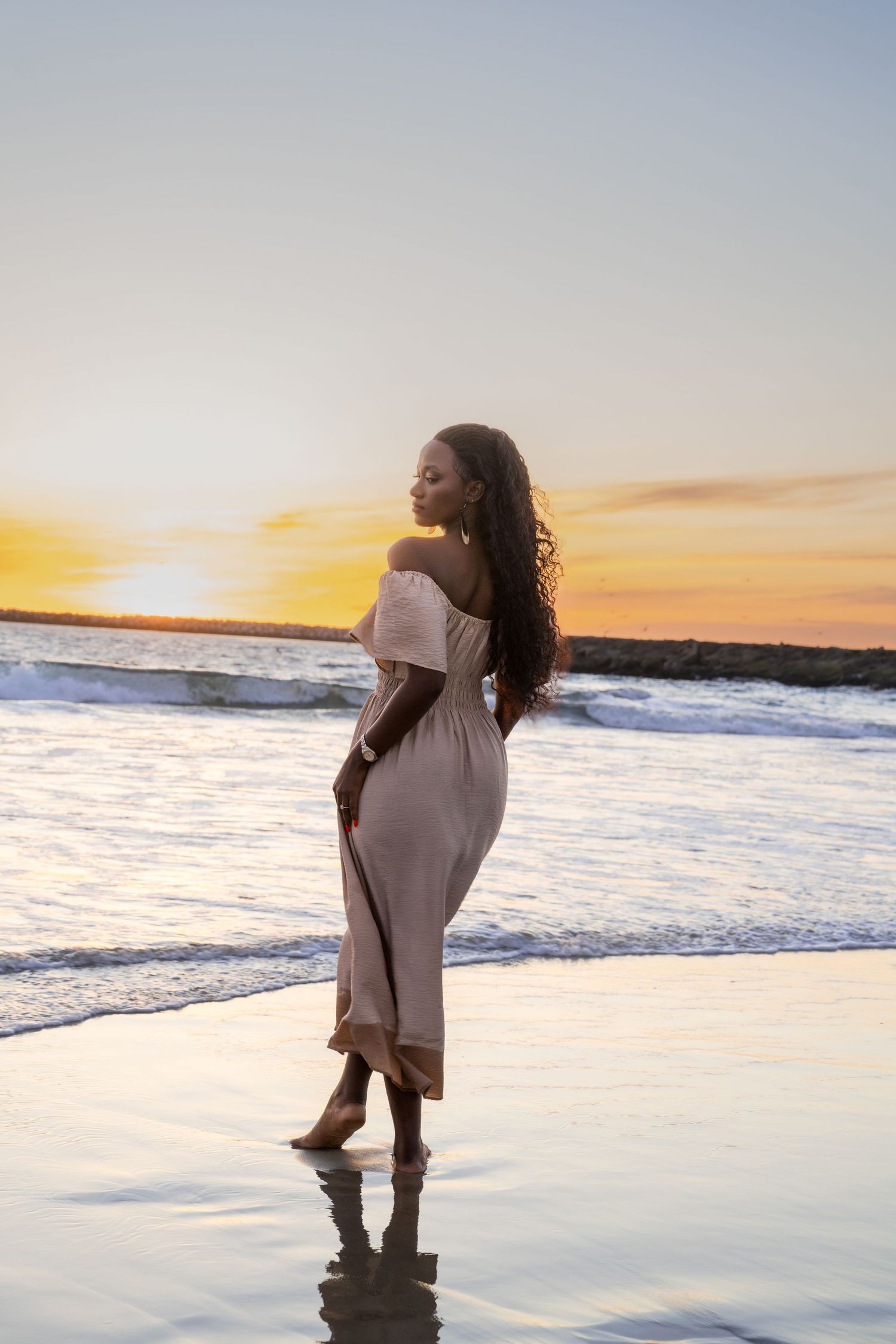 Silhouette of person in off-shoulder dress standing on beach at sunset.