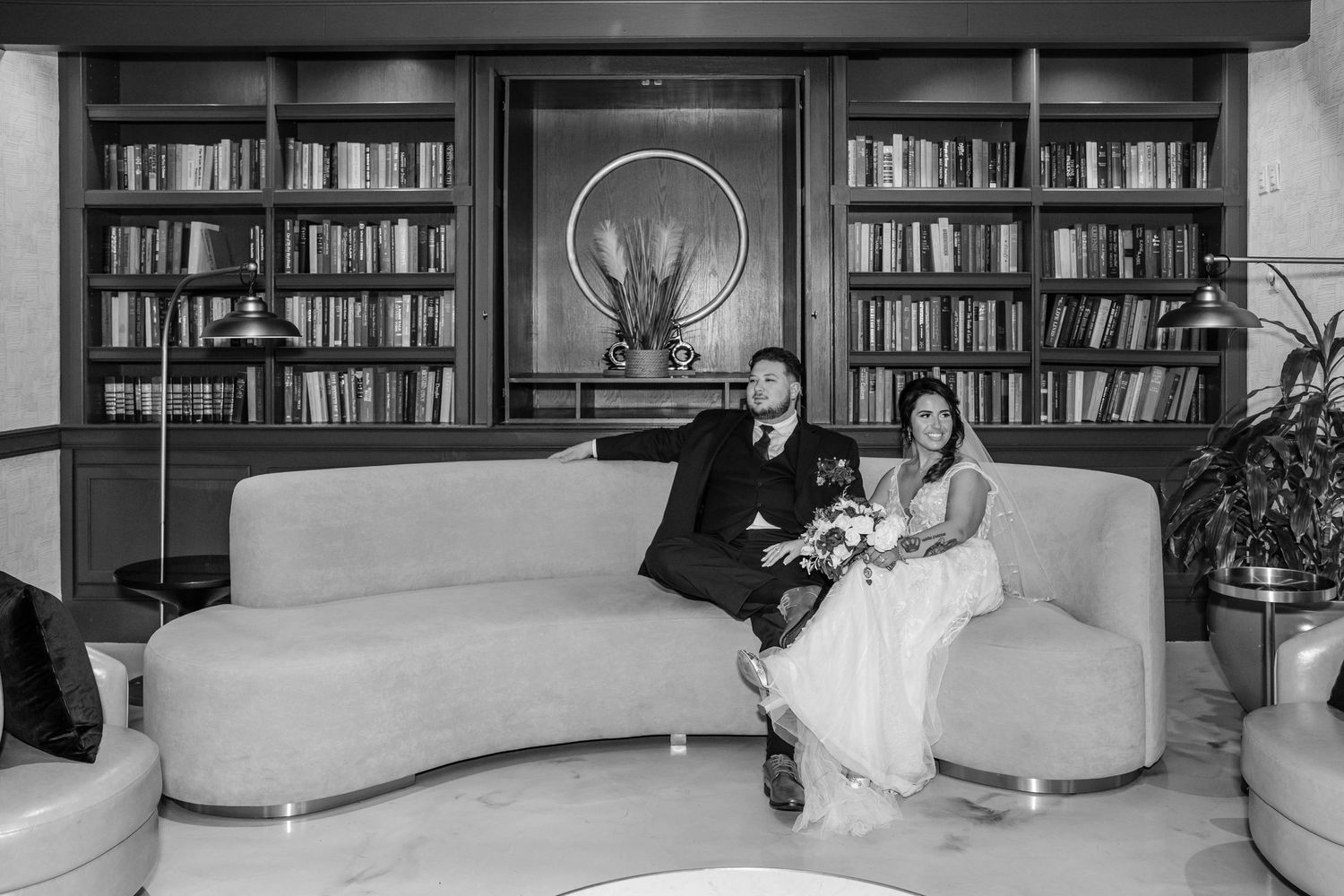 Black and white photo of couple seated on modern couch with library bookshelves behind them.