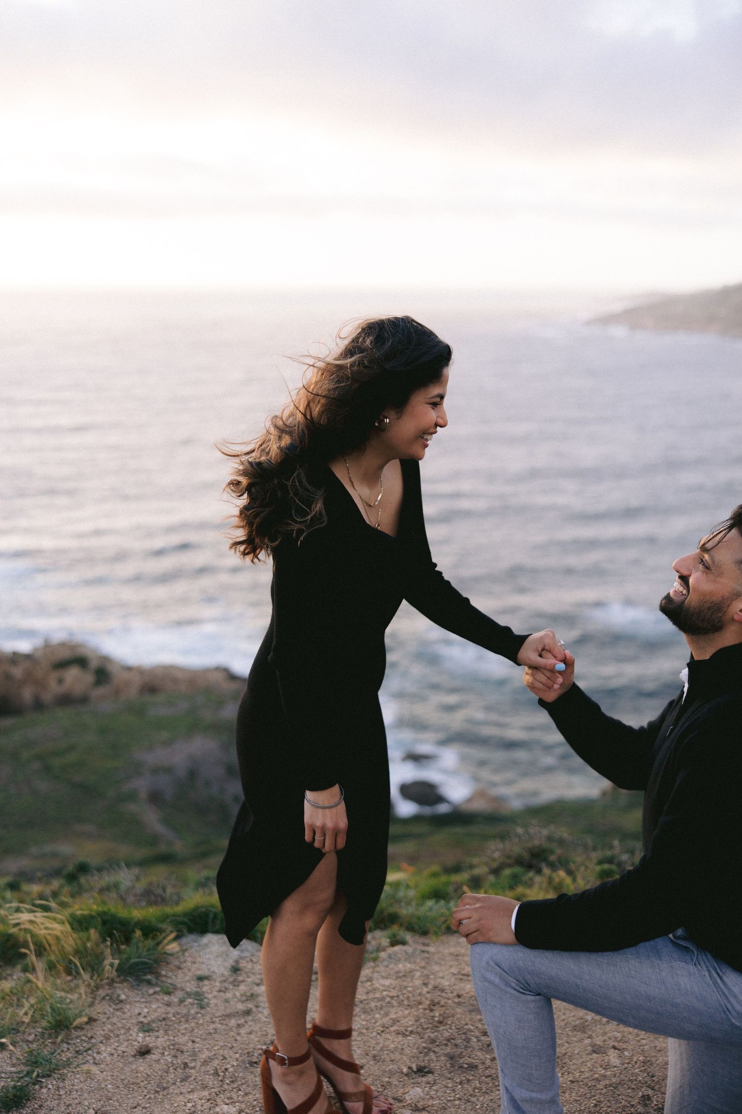 A romantic marriage proposal scene on a coastal cliff at sunset, with the ocean waves in the background.