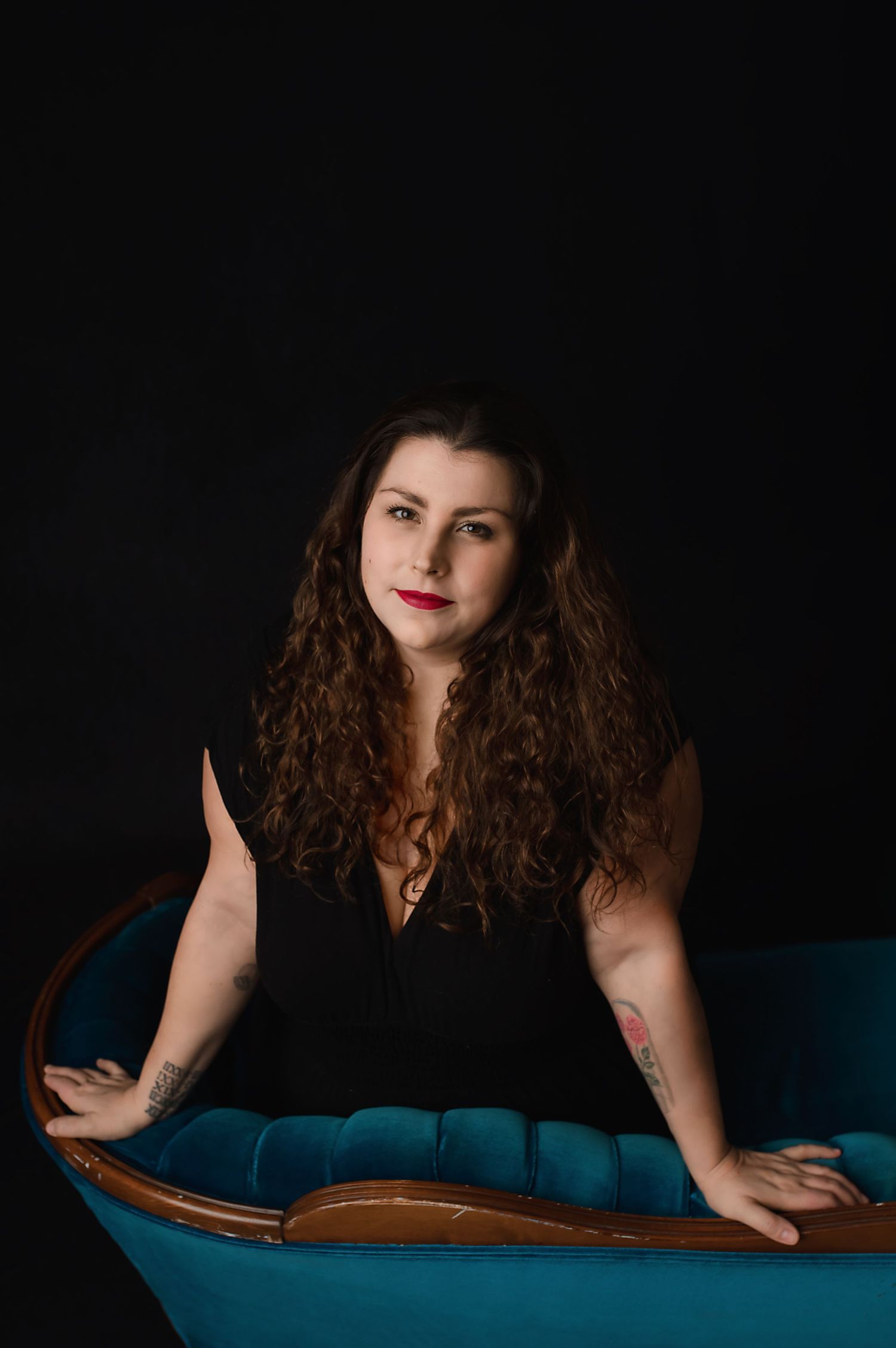 Artistic portrait of someone with long curly hair wearing a black sleeveless top seated on a teal blue chair against dark backdrop.