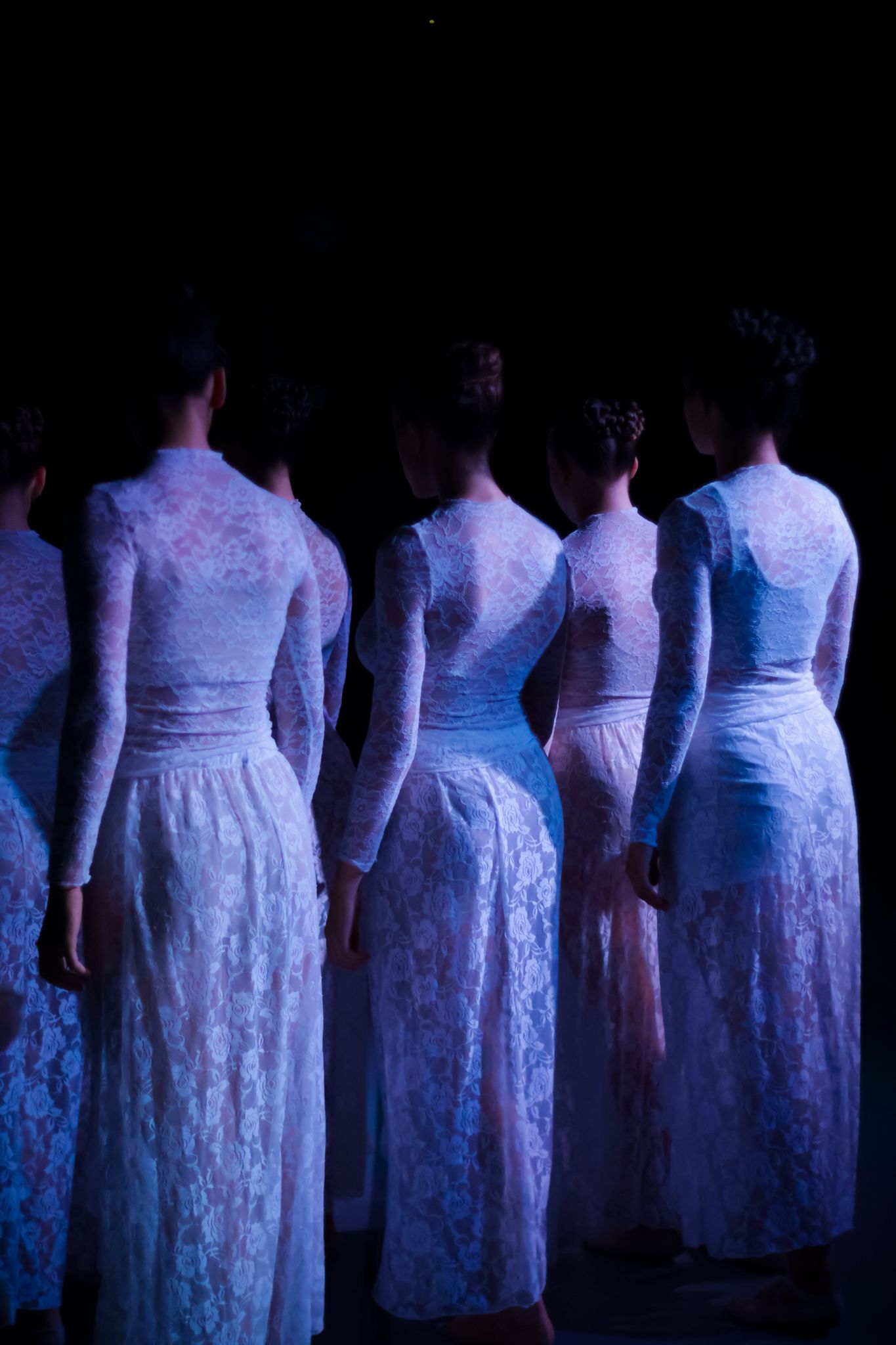 Row of performers in white dresses standing in dramatic blue stage lighting.