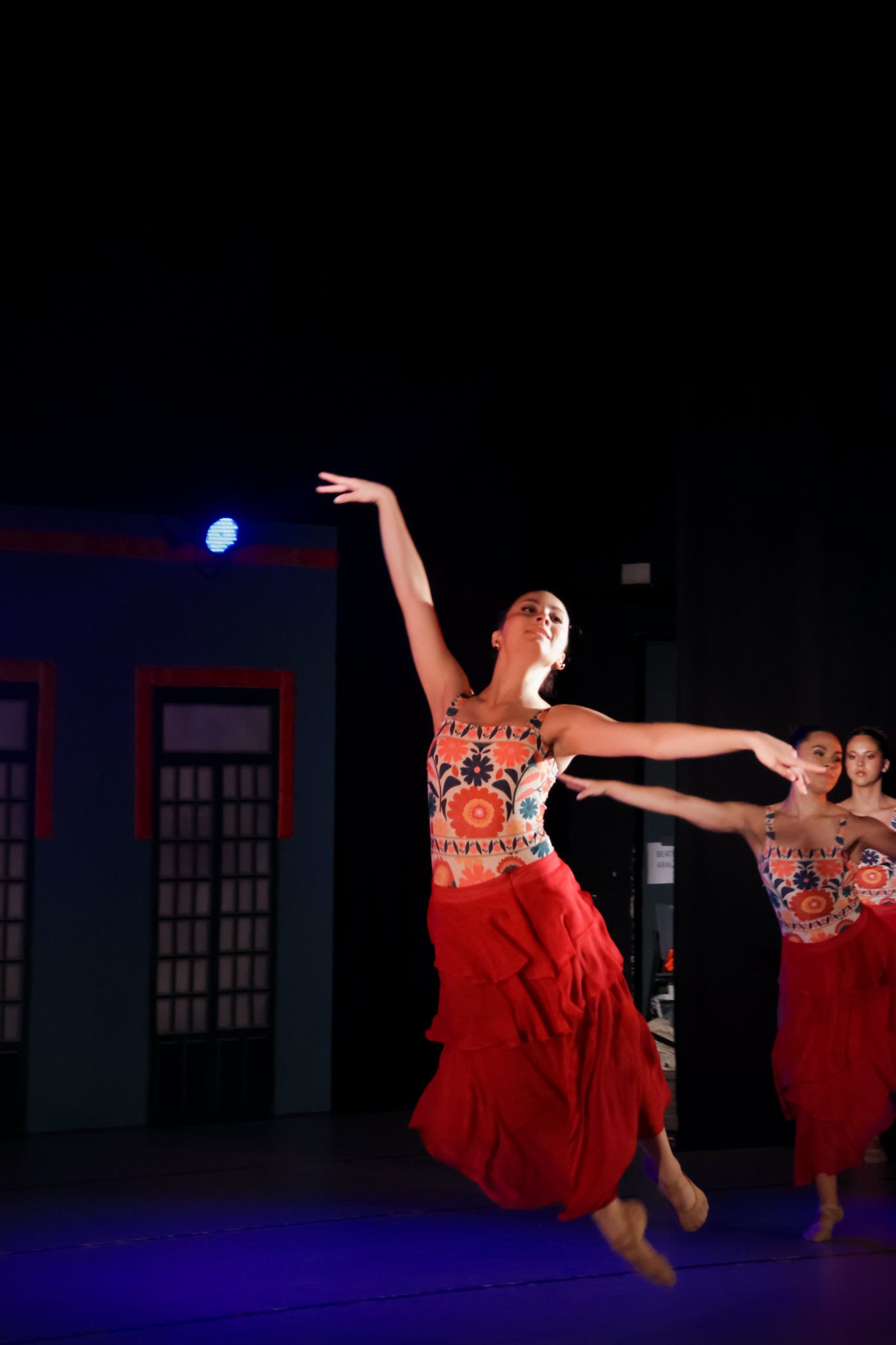 Dancers in flowing red costumes performing dynamic leaps on a dark stage.