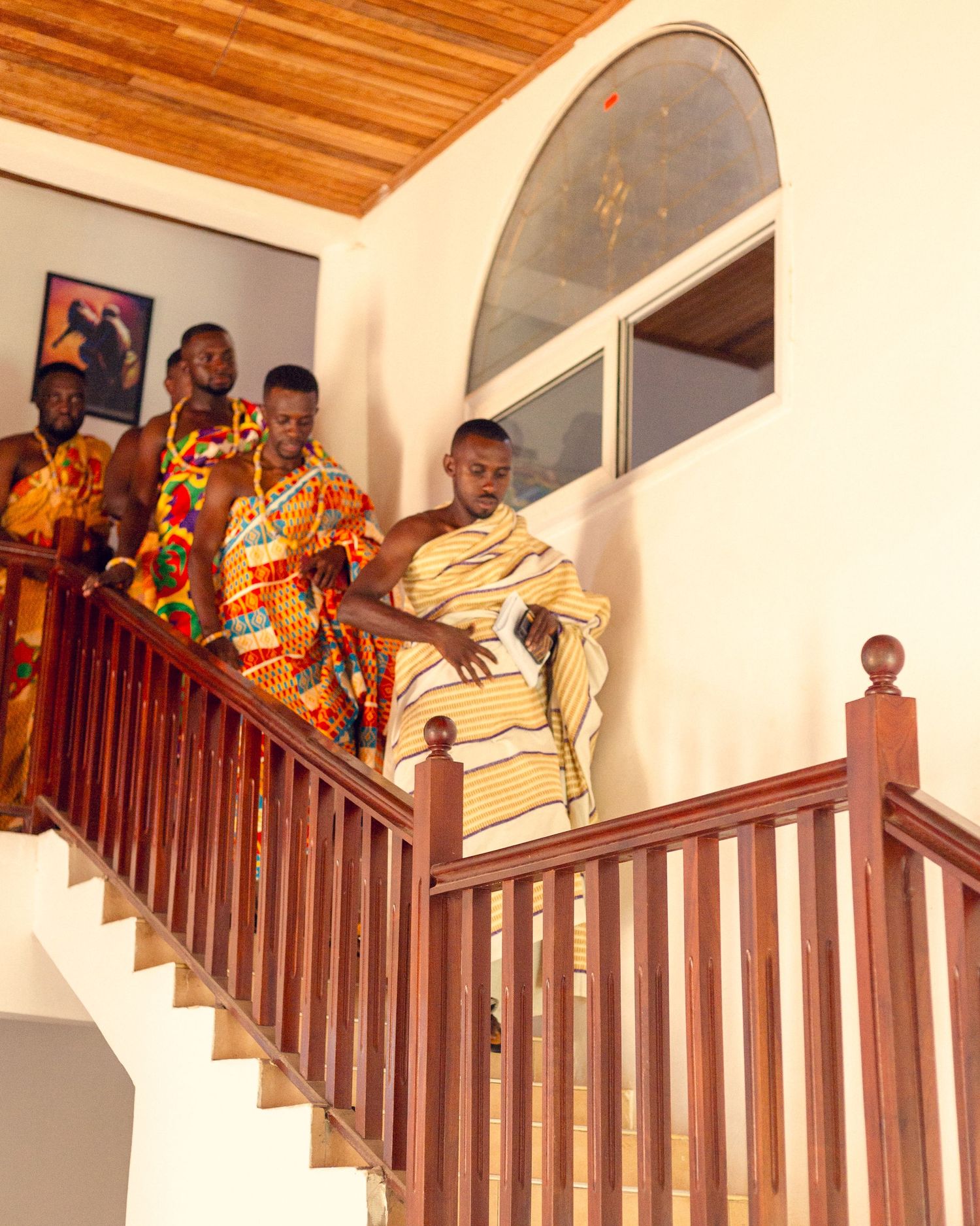Group wearing colorful African-inspired outfits pose on wooden staircase in bright interior setting.