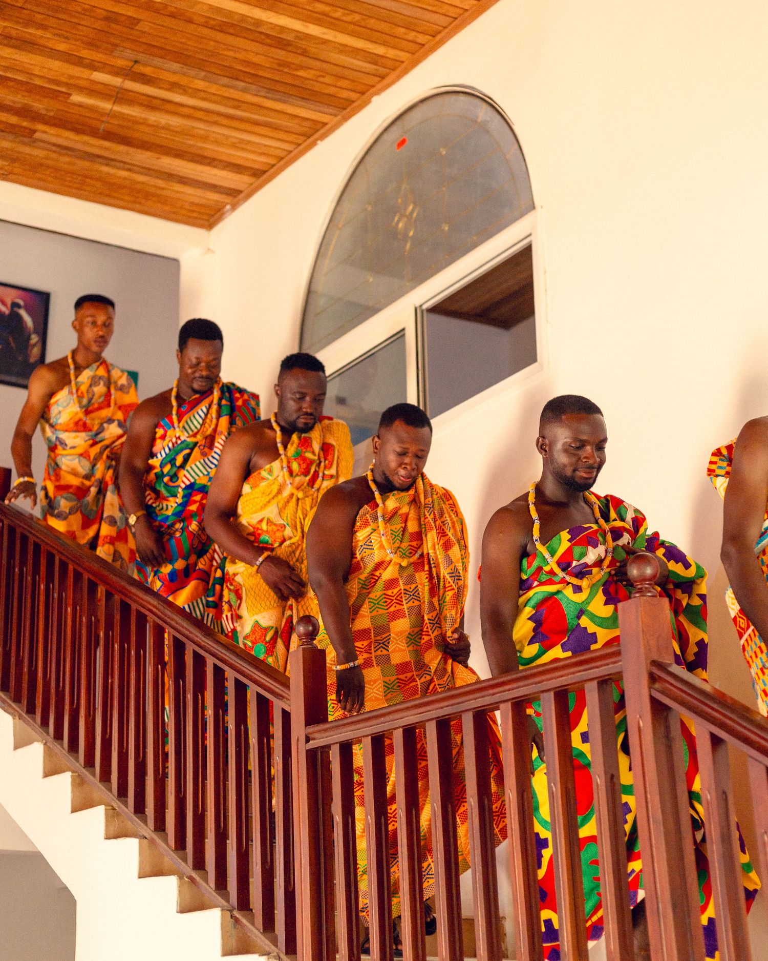 A group wearing colorful traditional African kente cloth and formal attire pose on wooden stairs with a mahogany railing.