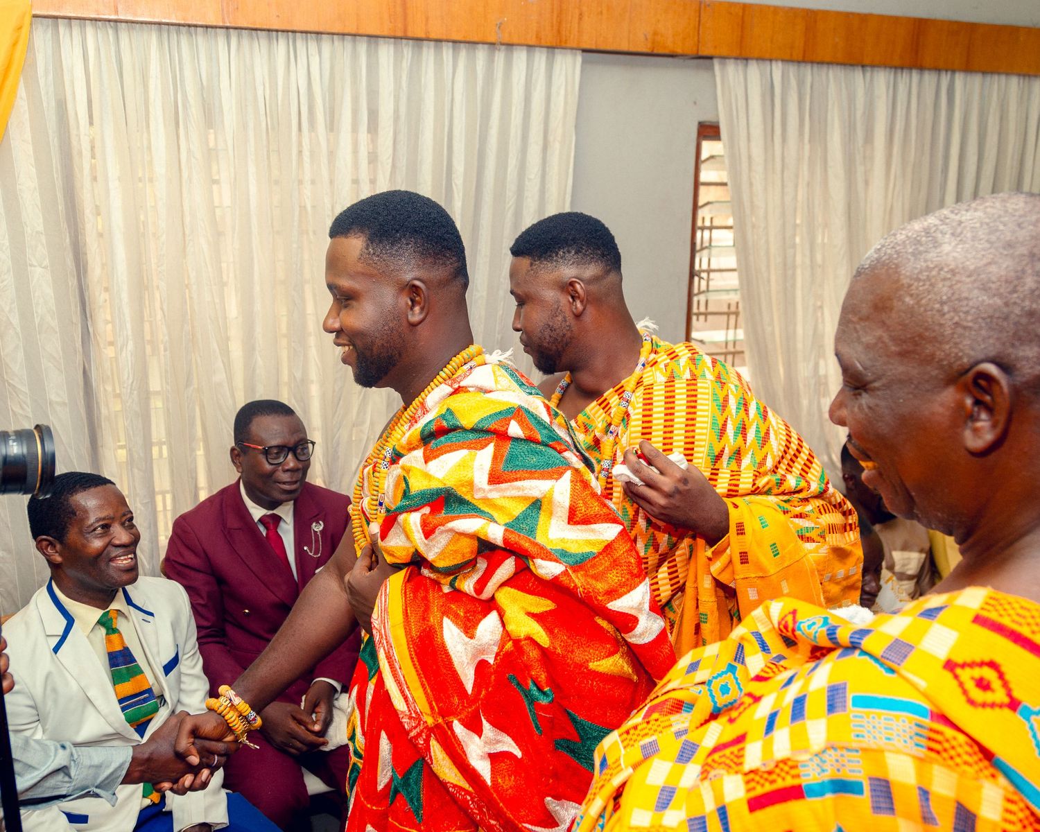 A group wearing vibrant traditional African kente cloth patterns and clothing gather indoors for an event.