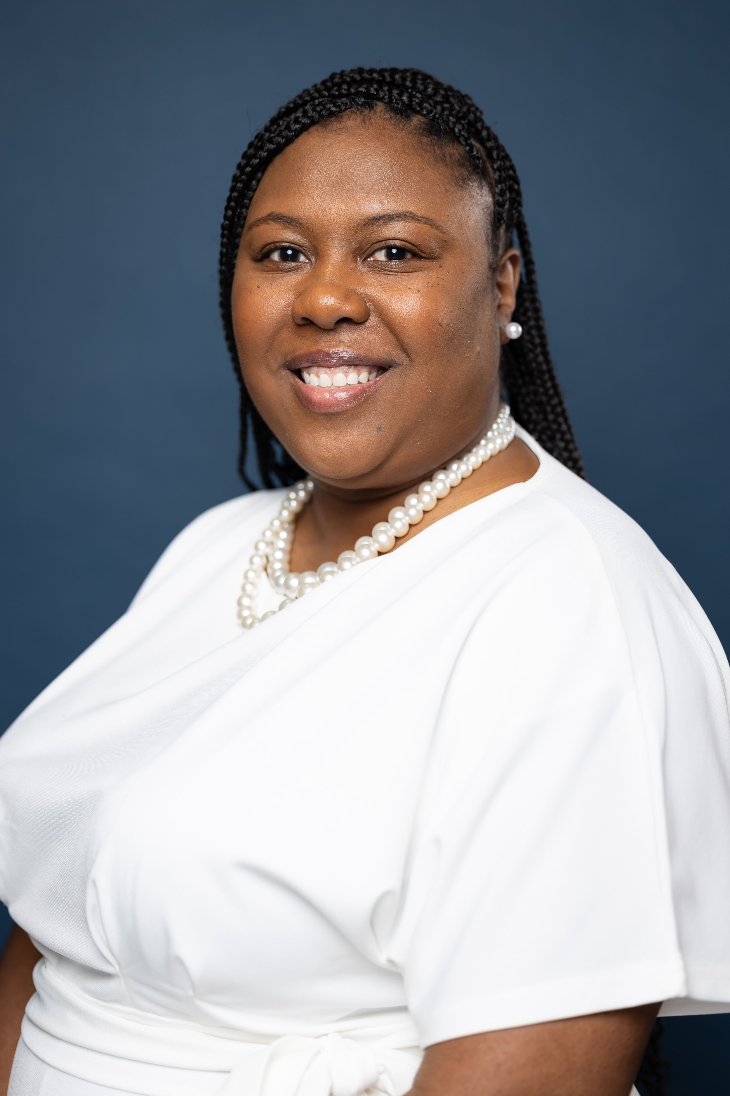 Professional portrait of someone in a white top with braided hair and pearl necklace against a blue background.