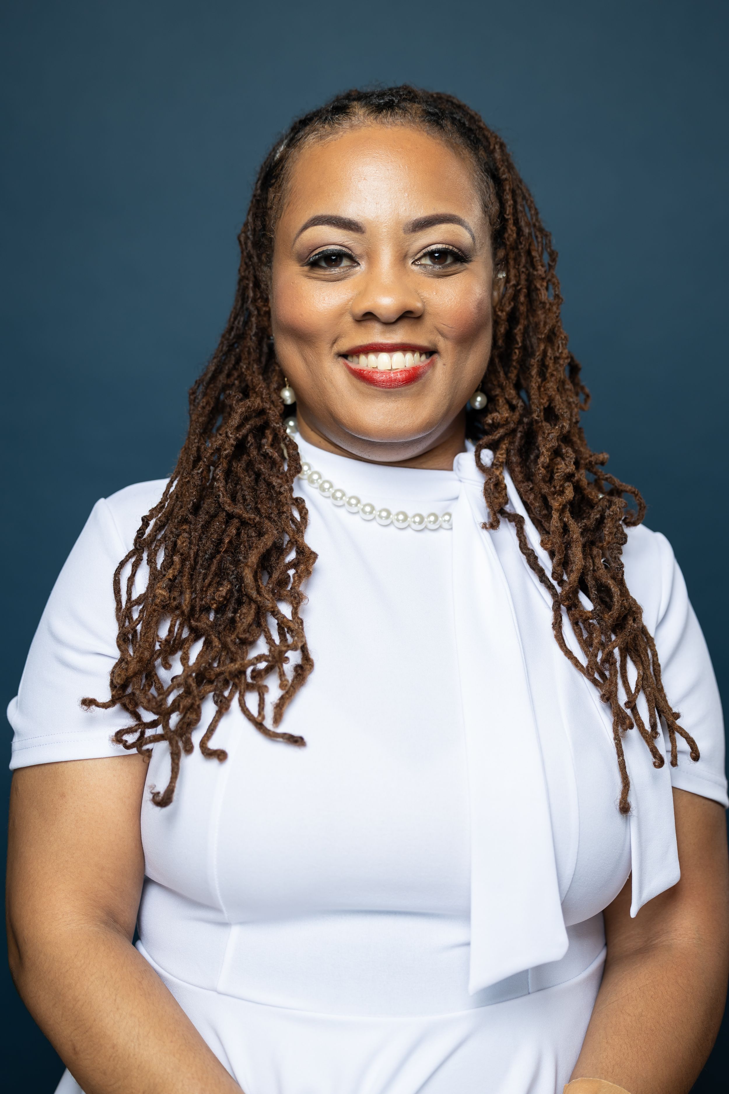 Professional headshot featuring long locs, white top and pearl necklace against dark backdrop.
