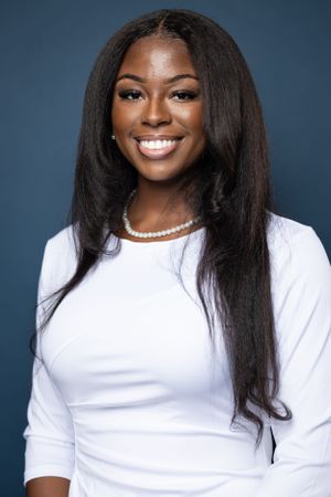 Professional portrait showing long straight hair, white top and pearl necklace against blue background.