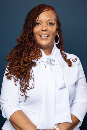 Studio portrait showcasing burgundy locs, white blazer and silver brooch against blue background.
