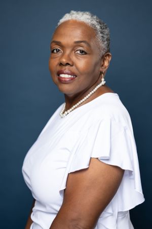 Professional headshot with short silver hair, pearl necklace and white top against dark backdrop.