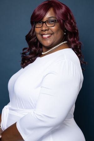 Professional portrait featuring burgundy waves, glasses and pearl necklace against blue background.