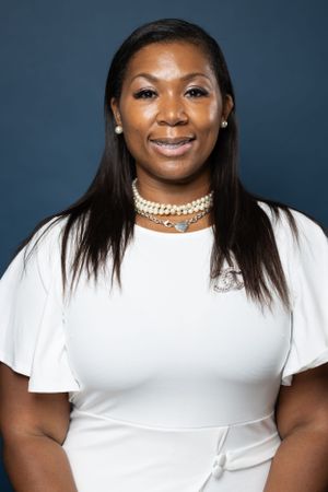 Professional headshot in white top with pearl choker necklace against dark blue background.