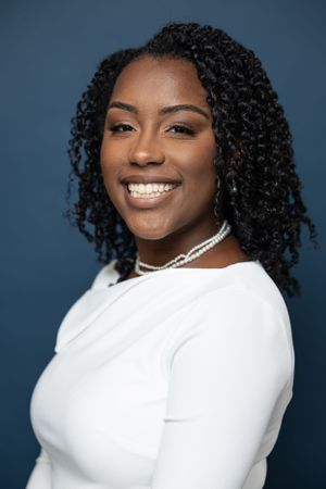 Professional portrait showing natural curls, white top and pearl necklace against blue backdrop.