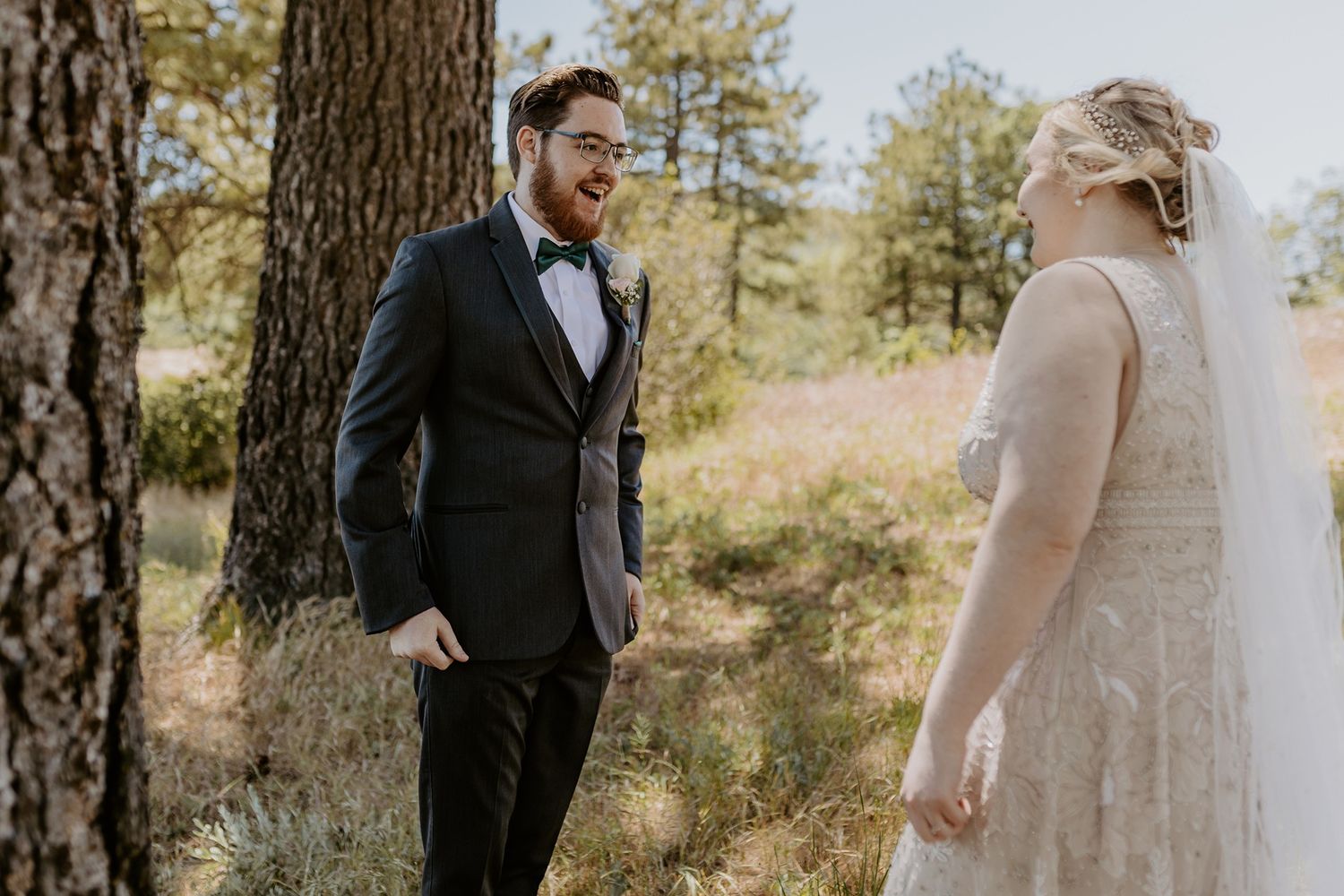 Groom in dark suit reacts joyfully seeing bride in lace gown during first look outdoors at Lake Cuyamaca, Julian.