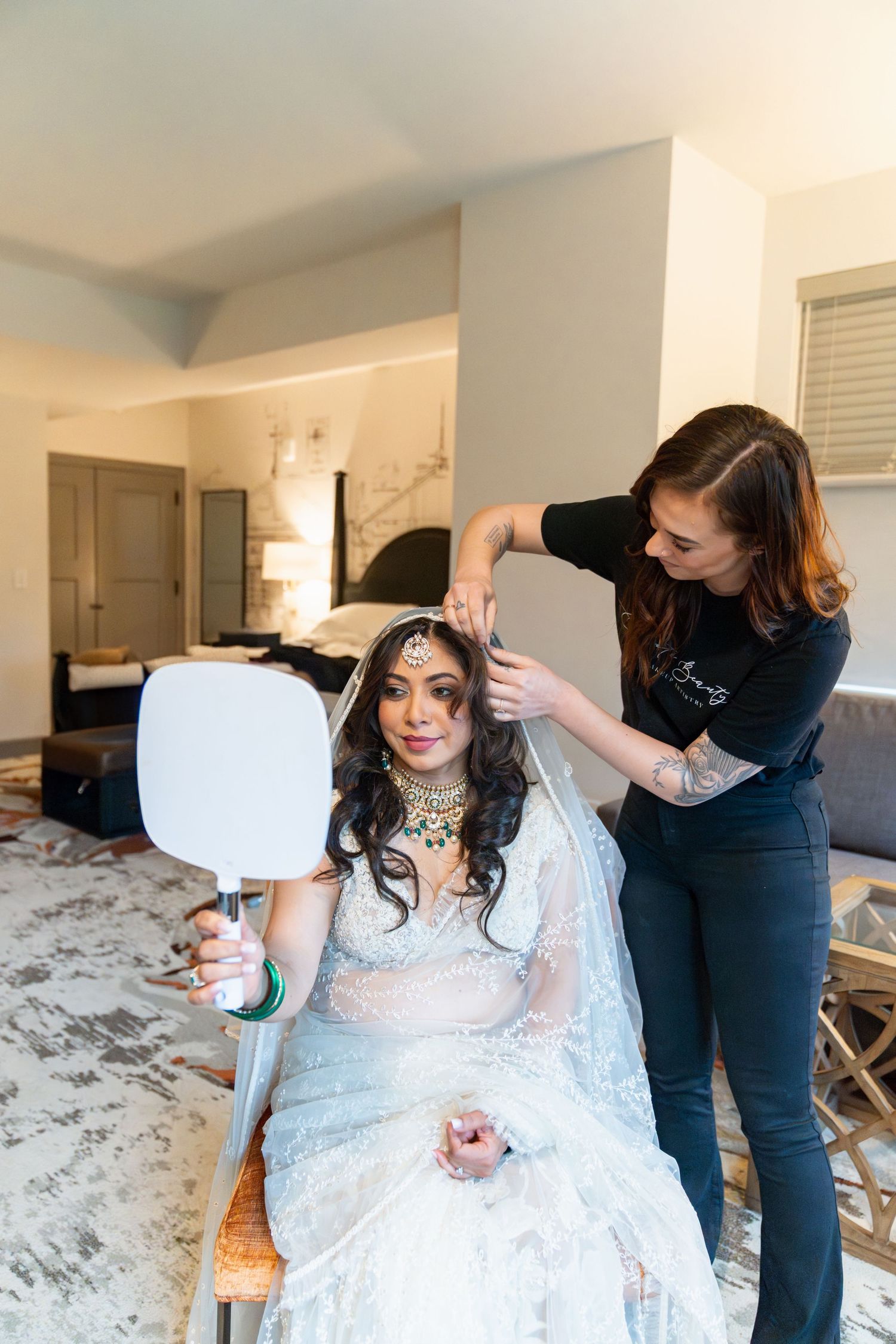 A makeup artist adjusts a veil on an Indian bride in a white lehenga holding a hand mirror during bridal preparations.