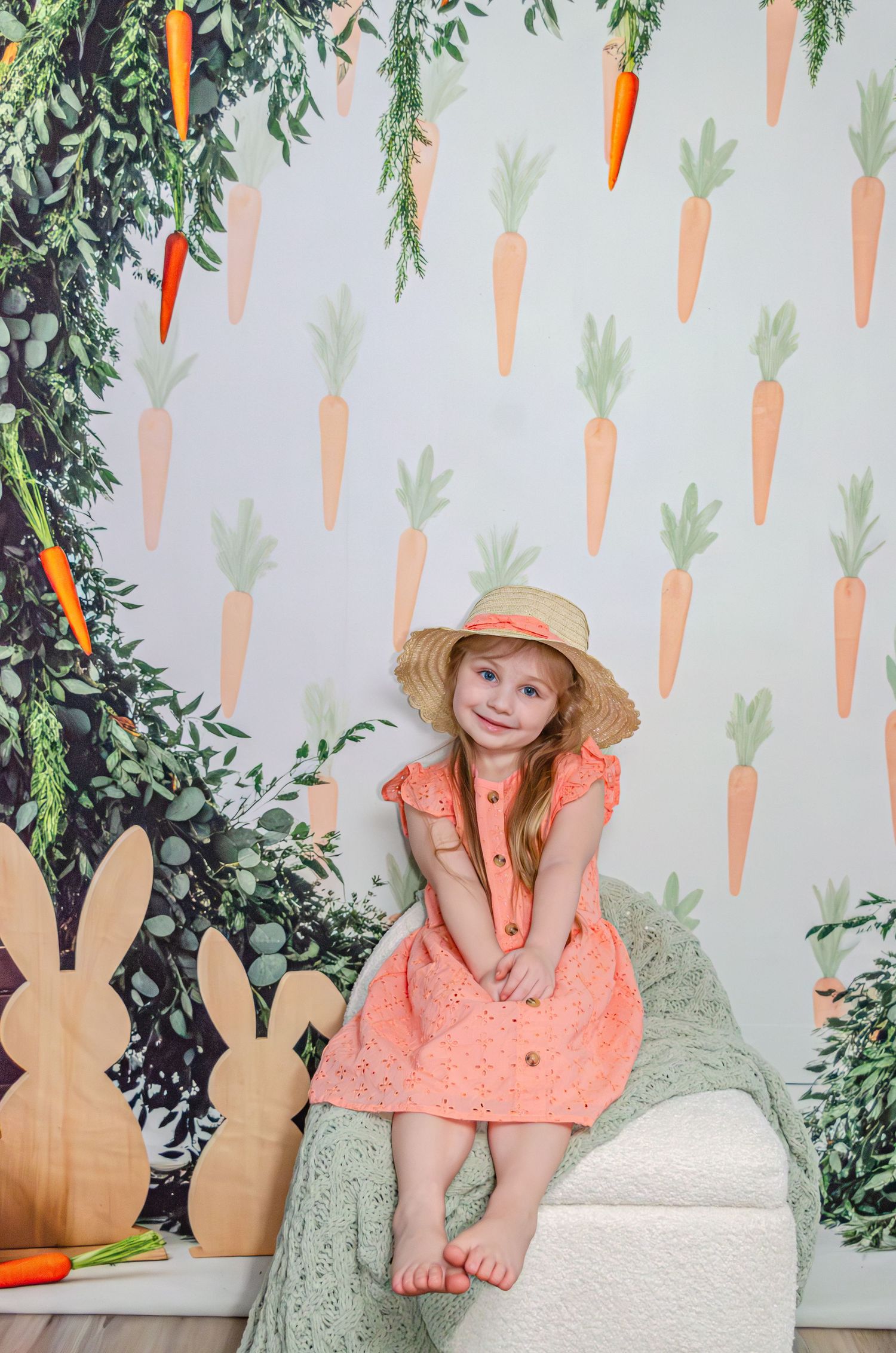 Baby in white dress with glasses sits in wooden crate beside FRAGILE box and lamp, Christmas tree background.