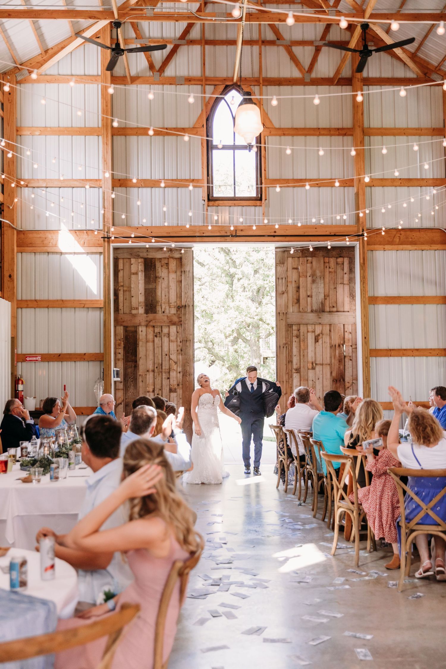 Bride and groom enter a rustic barn wedding reception with string lights, wooden doors, and guests seated at decorated tables.