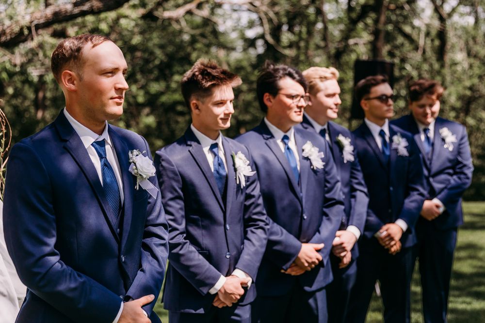 Groomsmen in navy blue suits with boutonnieres stand in a row outdoors at a wedding ceremony.