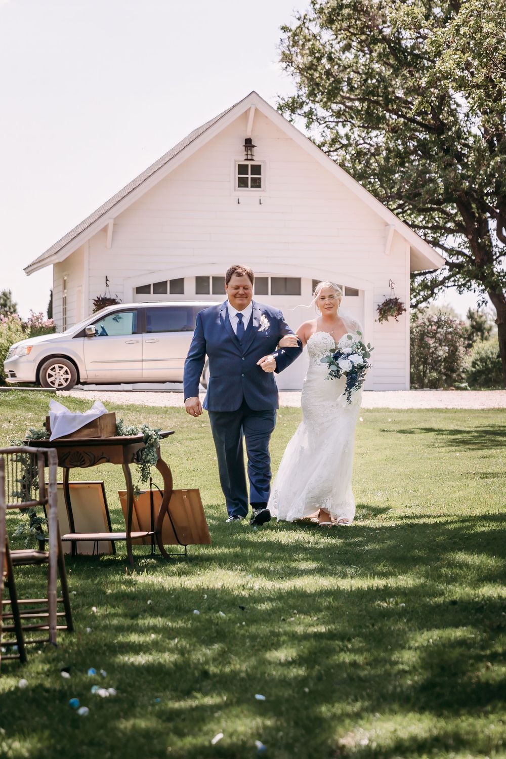 Bride and groom walking down outdoor aisle toward white barn venue on sunny wedding day.