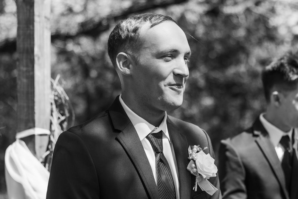 Smiling groom in black suit with boutonniere at outdoor wedding ceremony, black and white photo.