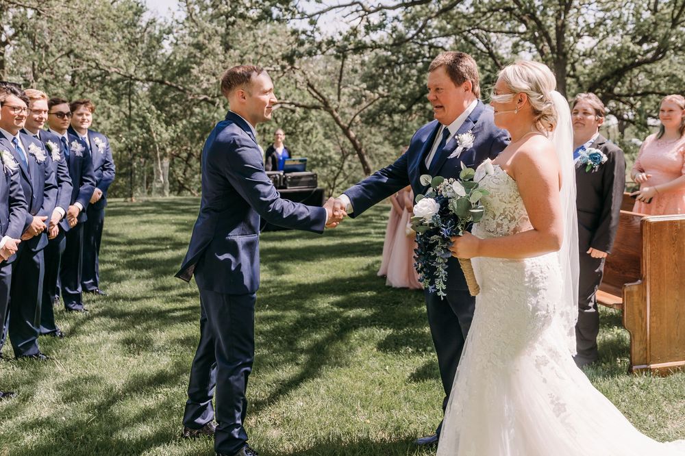 Groom shaking hands with bride's father during outdoor wedding ceremony with groomsmen lined up in navy suits.
