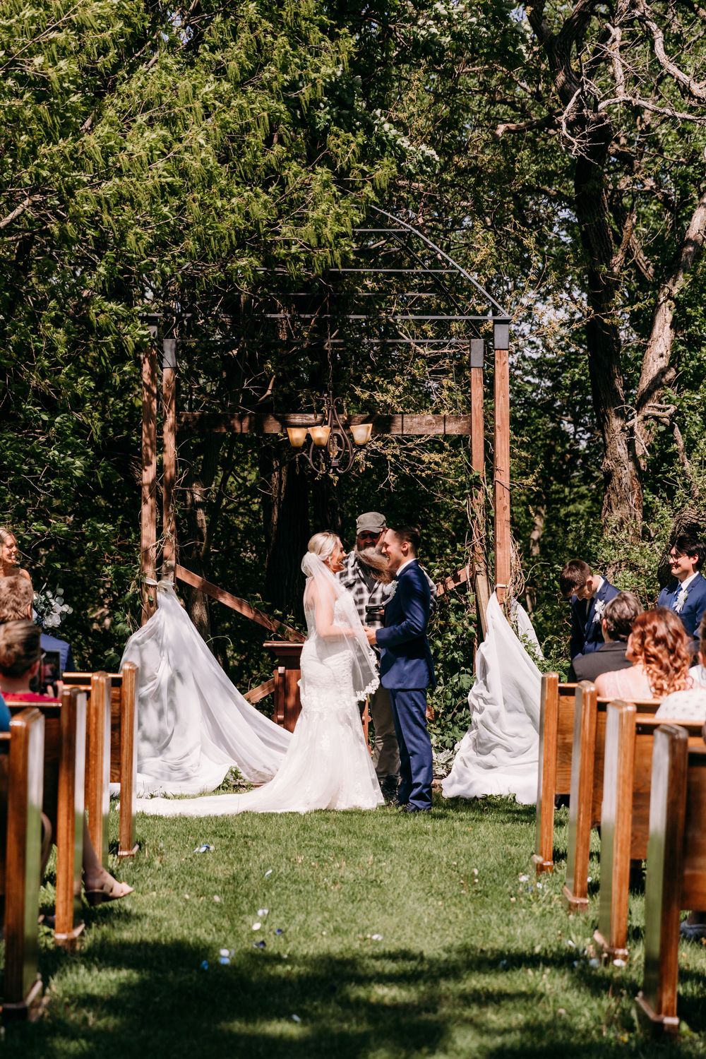Bride and groom exchange vows at an outdoor wedding ceremony under a wooden arch surrounded by lush green trees.