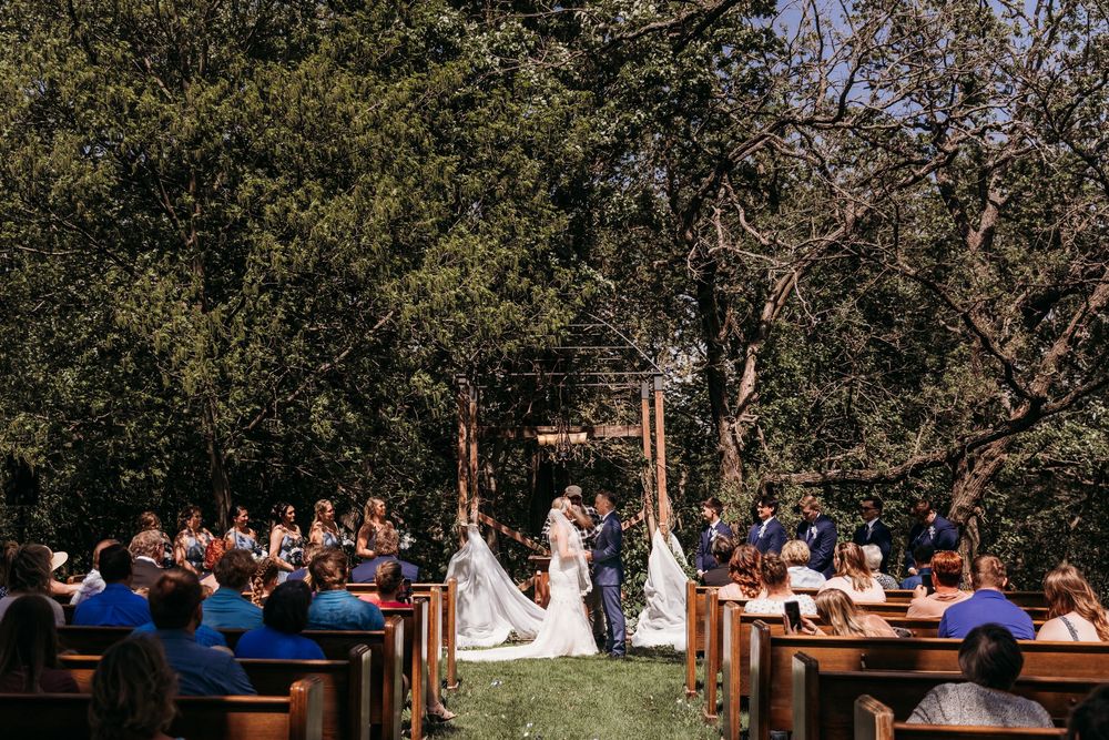 Outdoor wedding ceremony under trees with guests seated in wooden pews at rustic venue.
