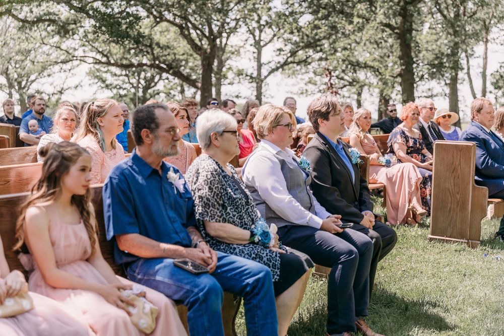 Guests seated on wooden benches at an outdoor wedding ceremony surrounded by trees on a sunny day.