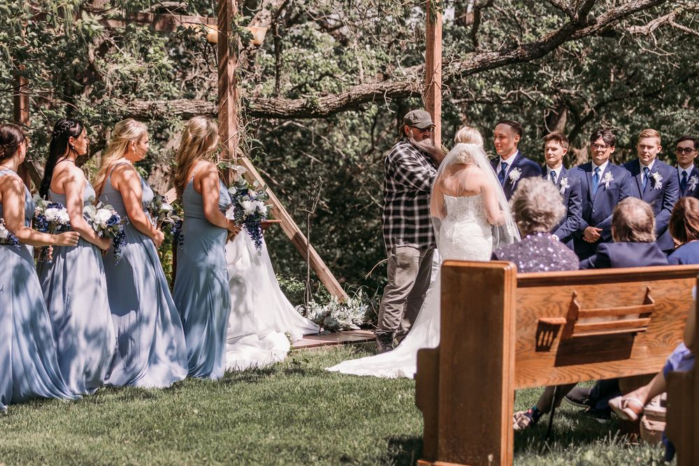 Outdoor wedding ceremony with bridesmaids in blue dresses standing beside bride and groom at rustic wooden altar.