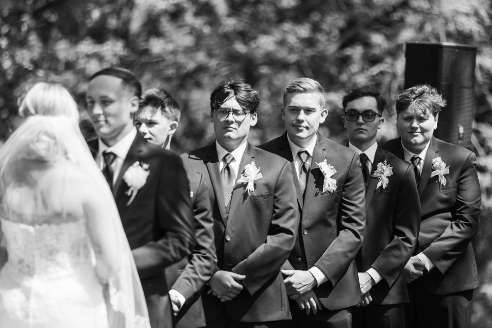 Groomsmen in dark suits with boutonnieres smile watching the bride walk down the aisle at an outdoor wedding.