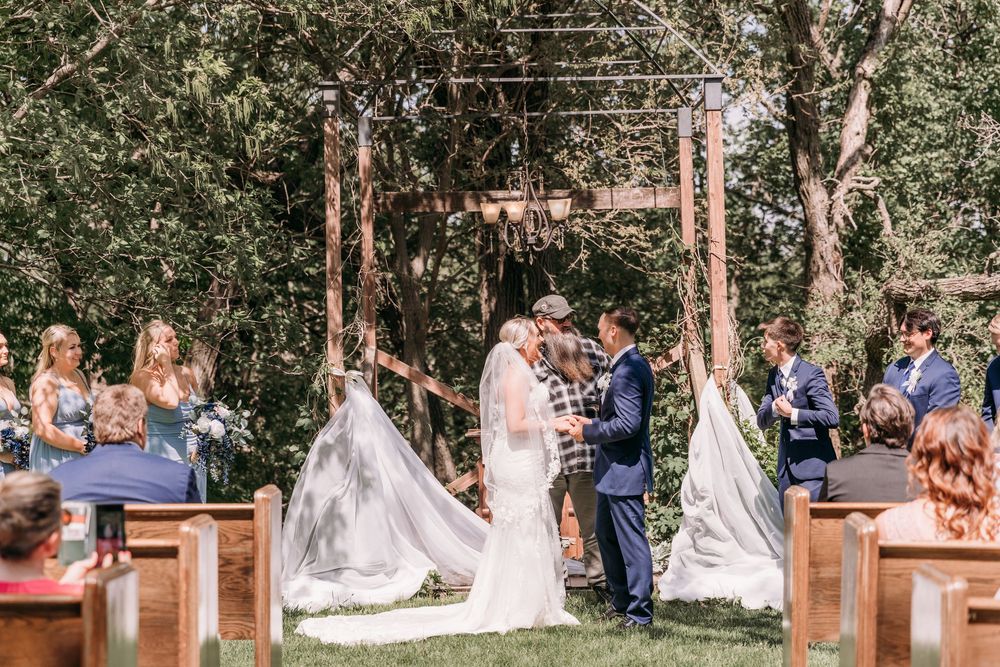 Outdoor wedding ceremony with bride and groom kissing under rustic wooden arch surrounded by lush trees and seated guests.