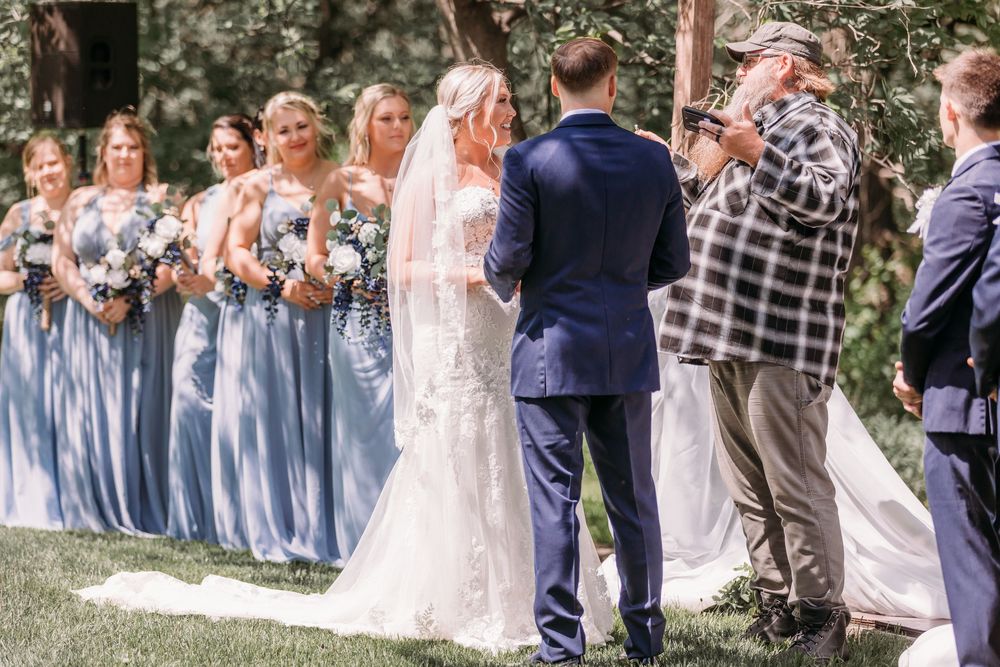 Bride in lace gown with veil stands at altar with groom surrounded by bridesmaids in blue dresses outdoors.