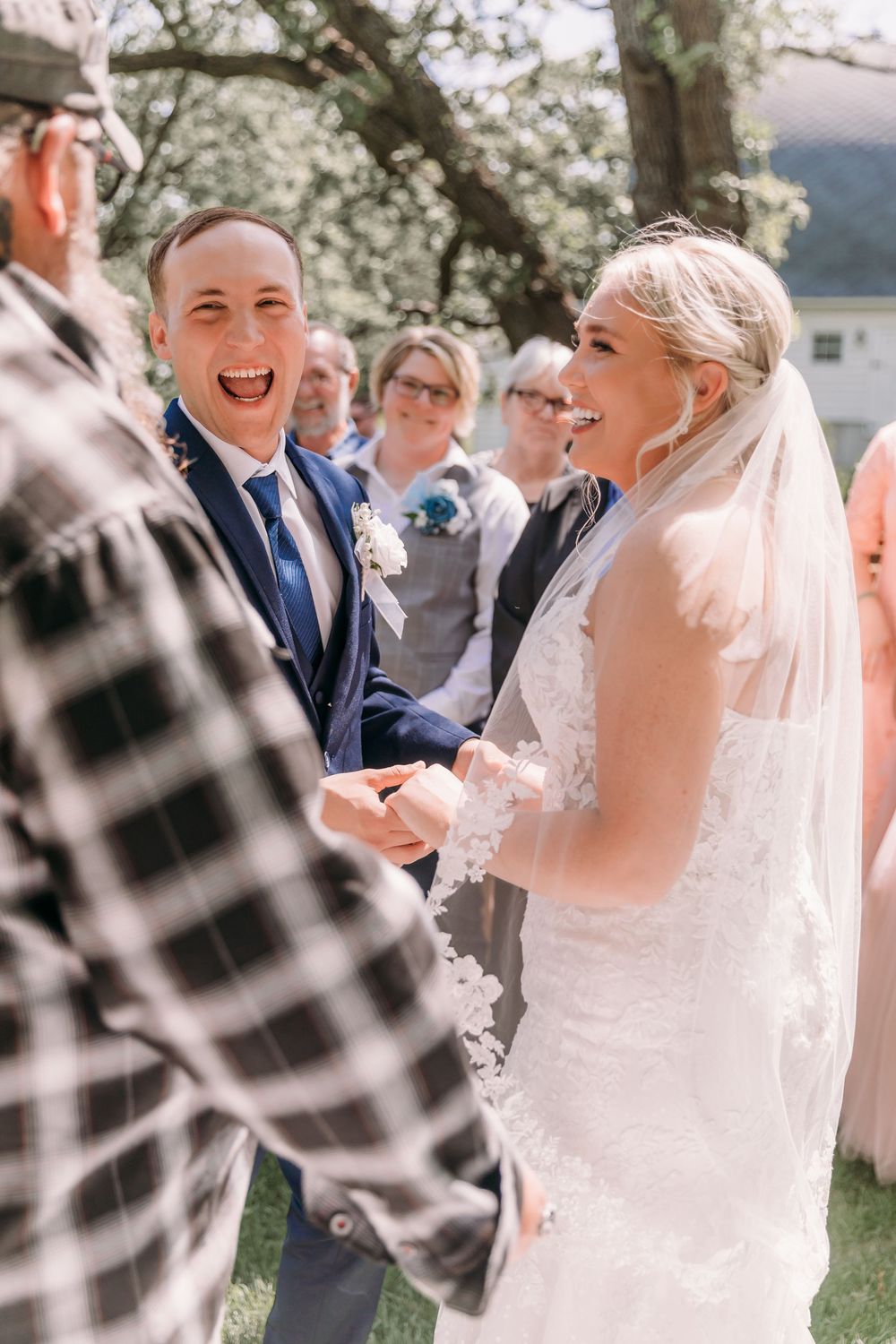 Joyful bride and groom laughing during outdoor wedding ceremony, bride in lace gown with veil, groom in navy suit.