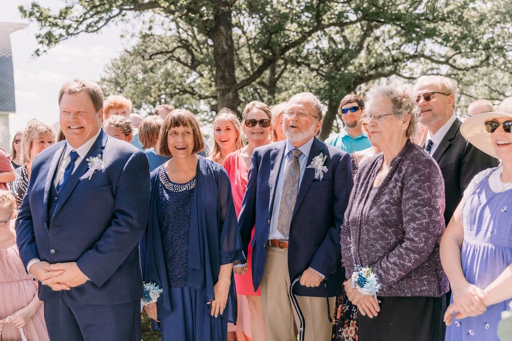 Wedding guests including family members smile and watch the outdoor ceremony on a sunny day.