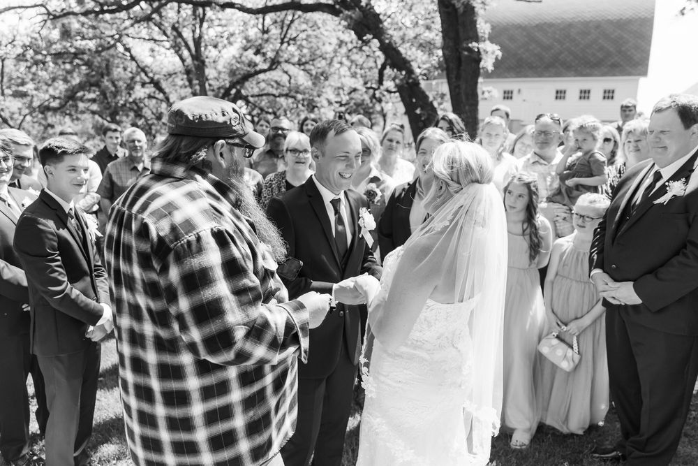 Black and white photo of bride and groom exchanging vows outdoors surrounded by wedding guests.