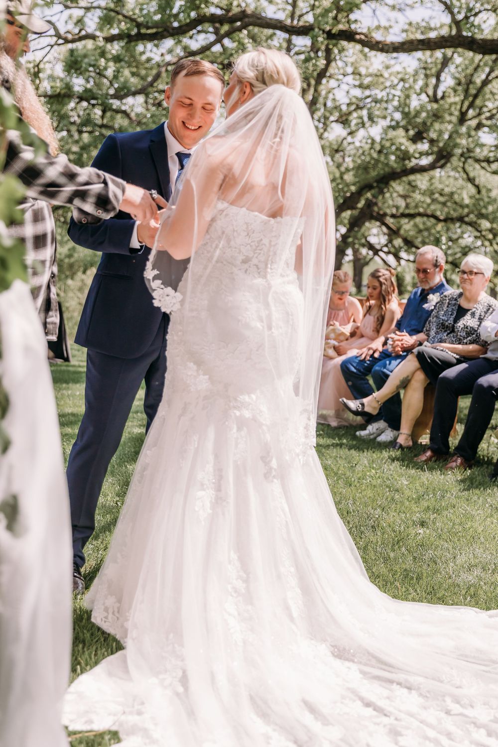 Smiling groom in navy suit places ring on bride in lace mermaid gown during outdoor wedding ceremony.