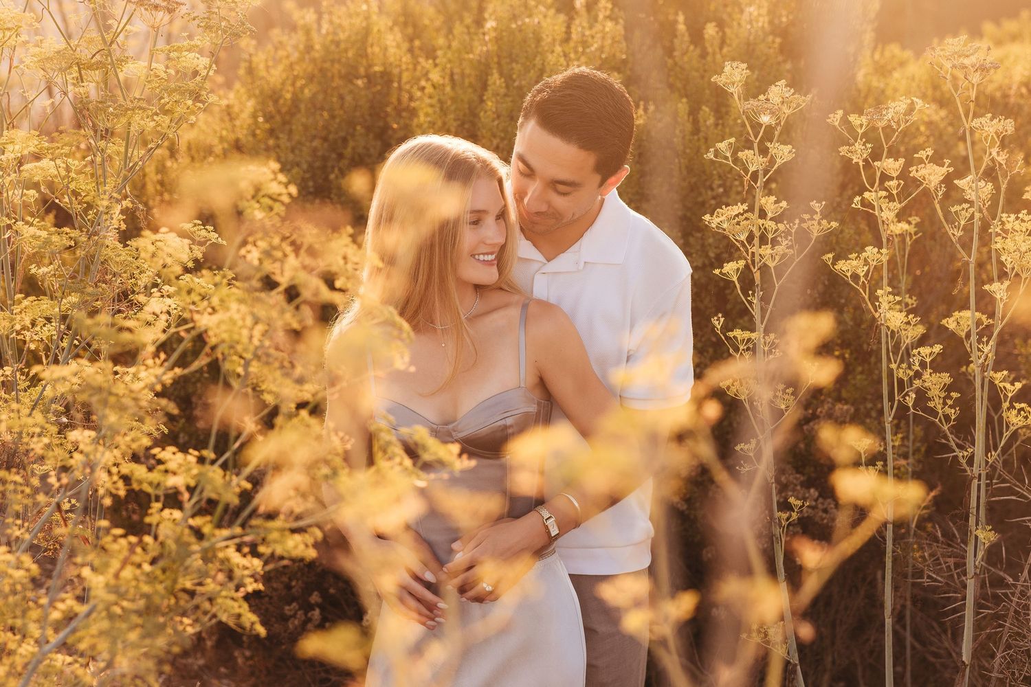 A couple embraces romantically in a golden sunlit field of yellow wildflowers during a warm sunset.