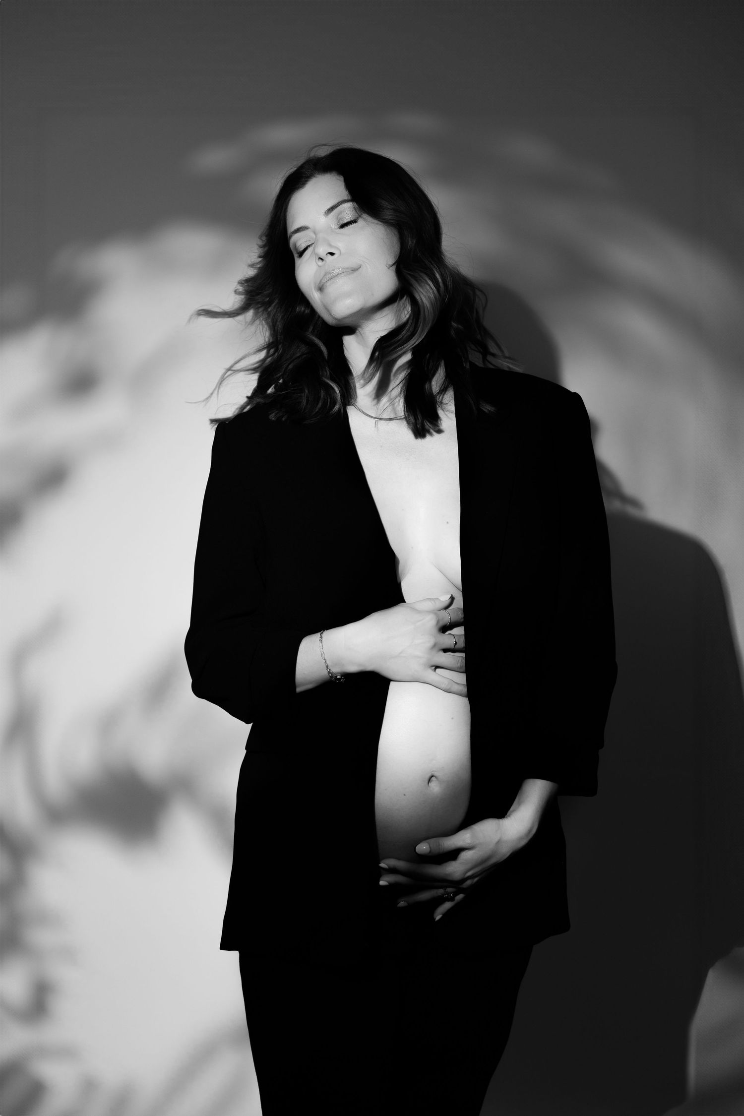 Pregnant woman in black off-shoulder gown poses gracefully with flowing beige fabric backdrop in elegant maternity photo.