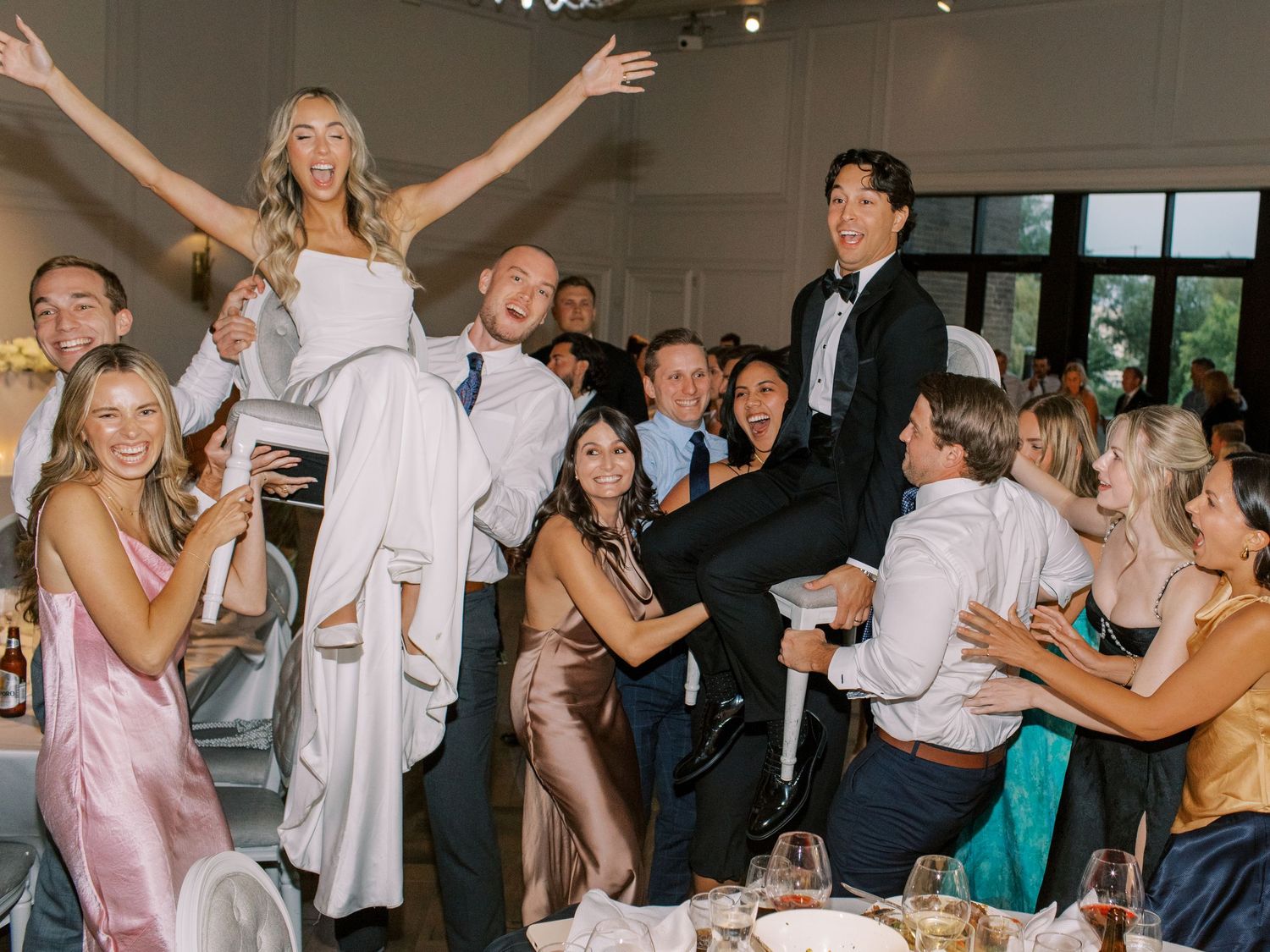 Joyful wedding reception guests lifting bride and groom in celebration at elegant indoor venue.